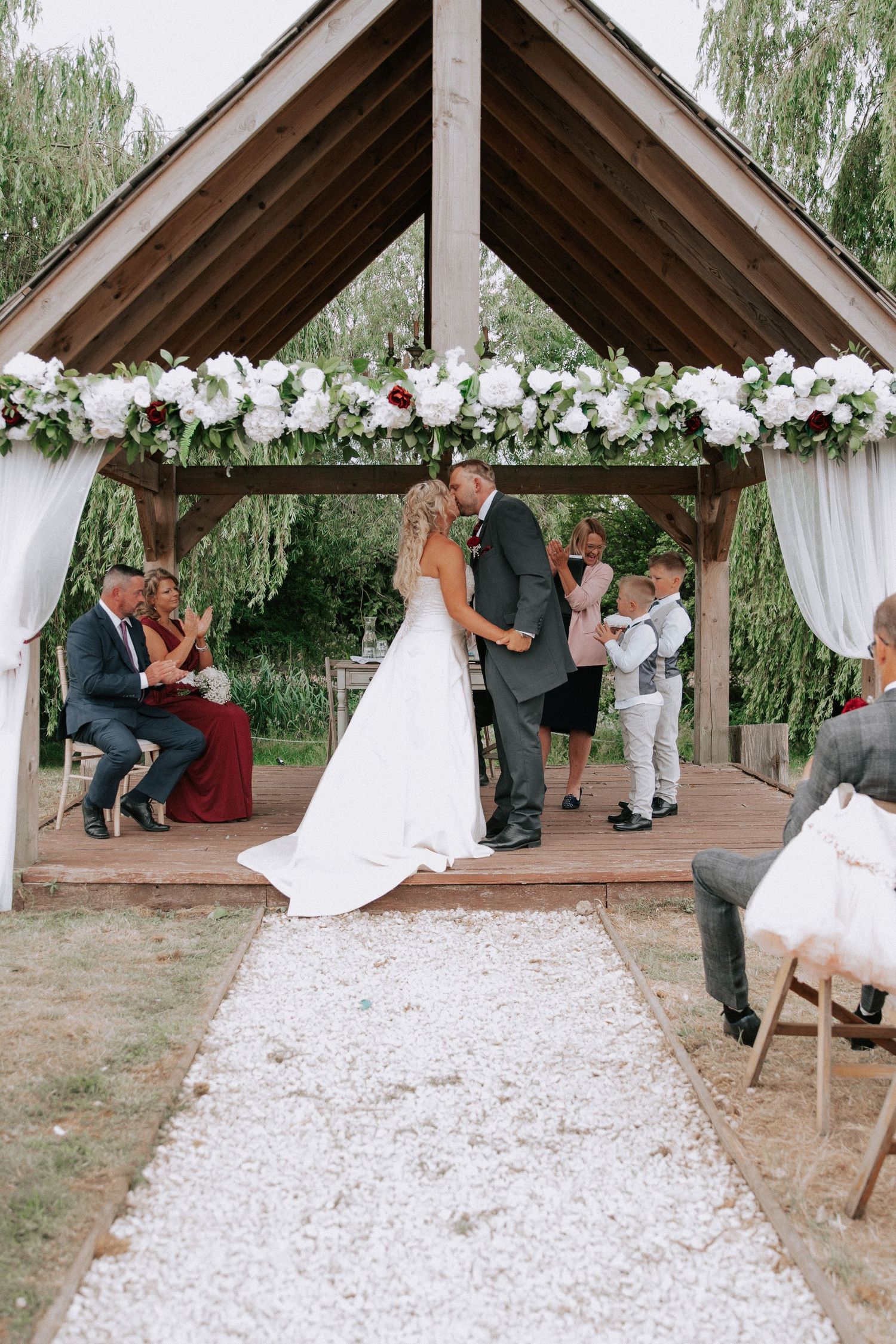 Bride and groom share first kiss under floral-adorned wooden gazebo during outdoor wedding ceremony with white petal aisle.