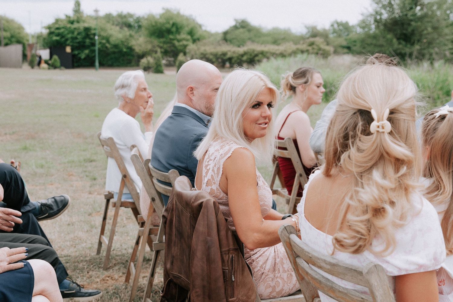 Guests seated on wooden chairs at an outdoor wedding ceremony, with lush greenery in the background.