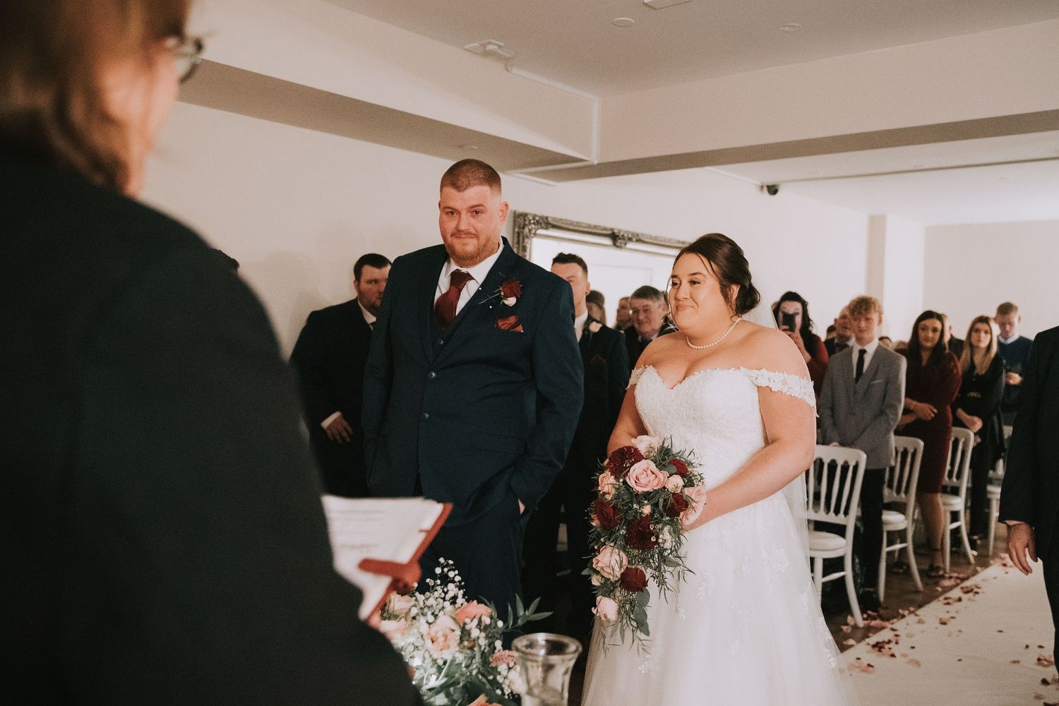Bride and groom stand at the altar during their wedding ceremony, bride holding a bouquet of pink and burgundy flowers.