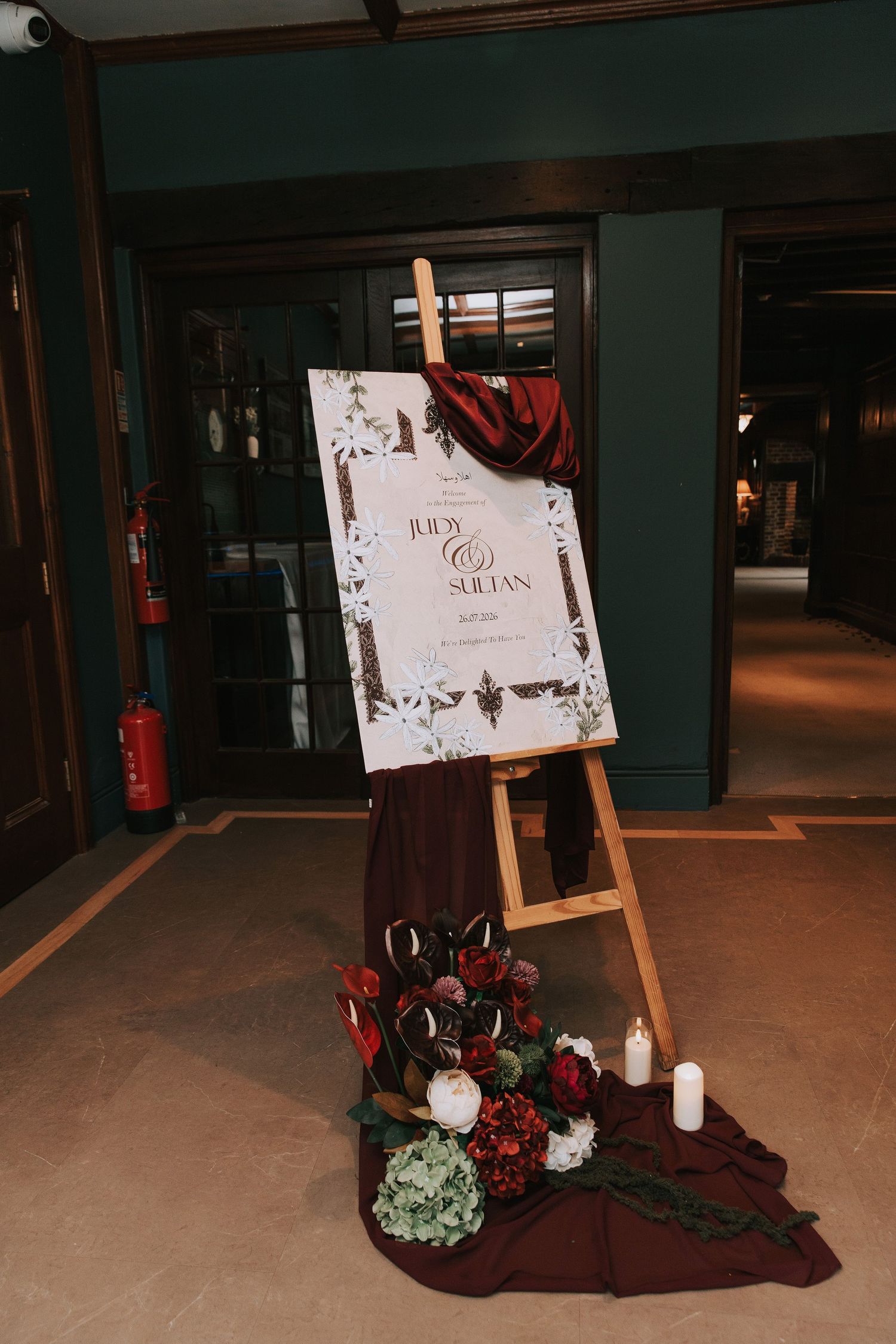 Elegant wedding welcome sign on wooden easel with burgundy drape, florals, and candles in a dimly lit venue.