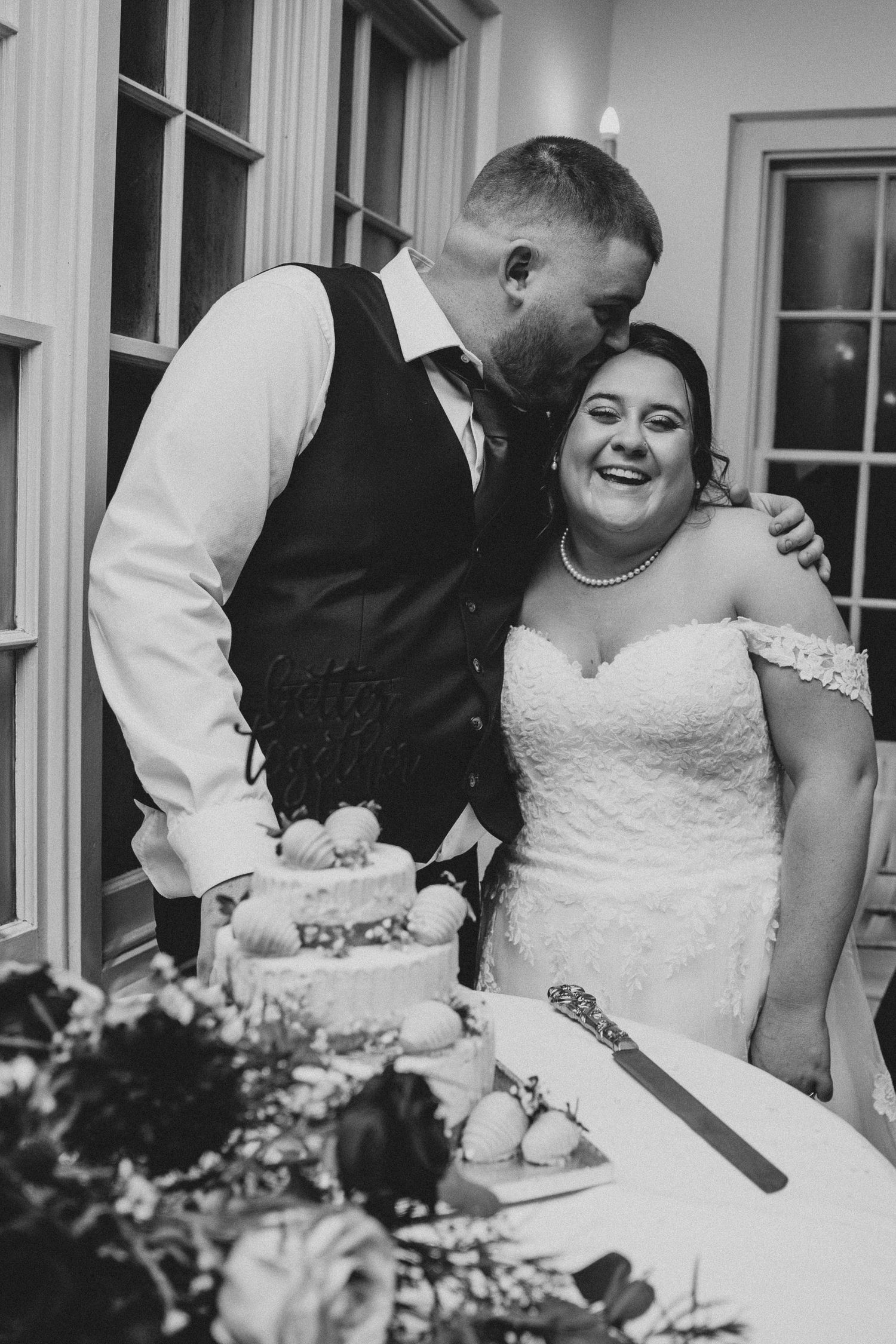 Black and white photo of smiling bride and groom sharing a kiss while cutting their wedding cake together.