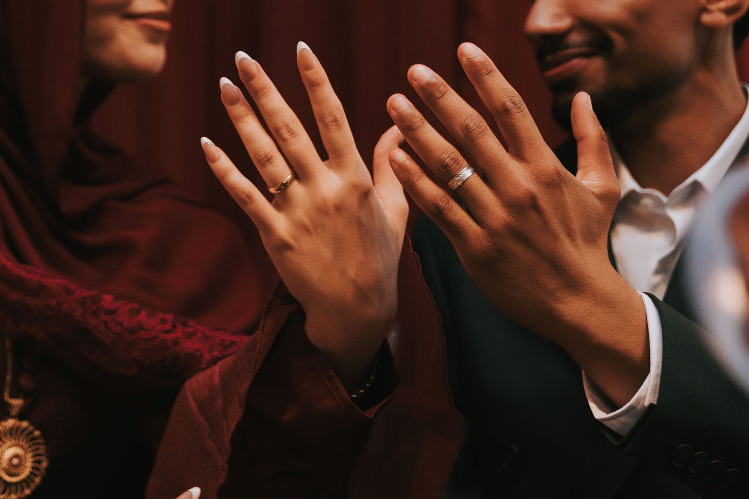 Couple displaying wedding rings on raised hands, woman in red hijab and man in suit, celebrating marriage.