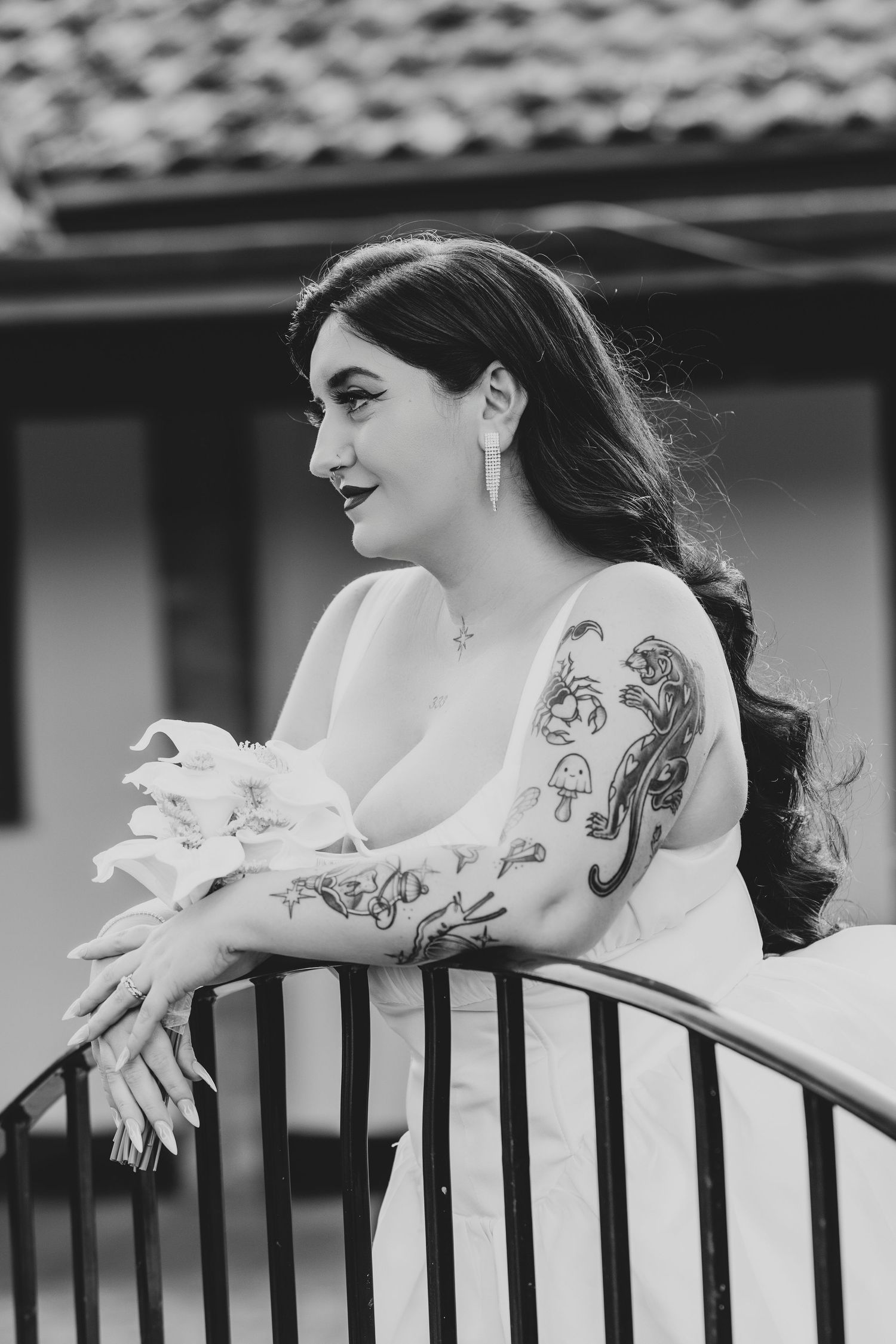 Black and white portrait of a tattooed bride in white dress leaning on iron railing, holding flowers.