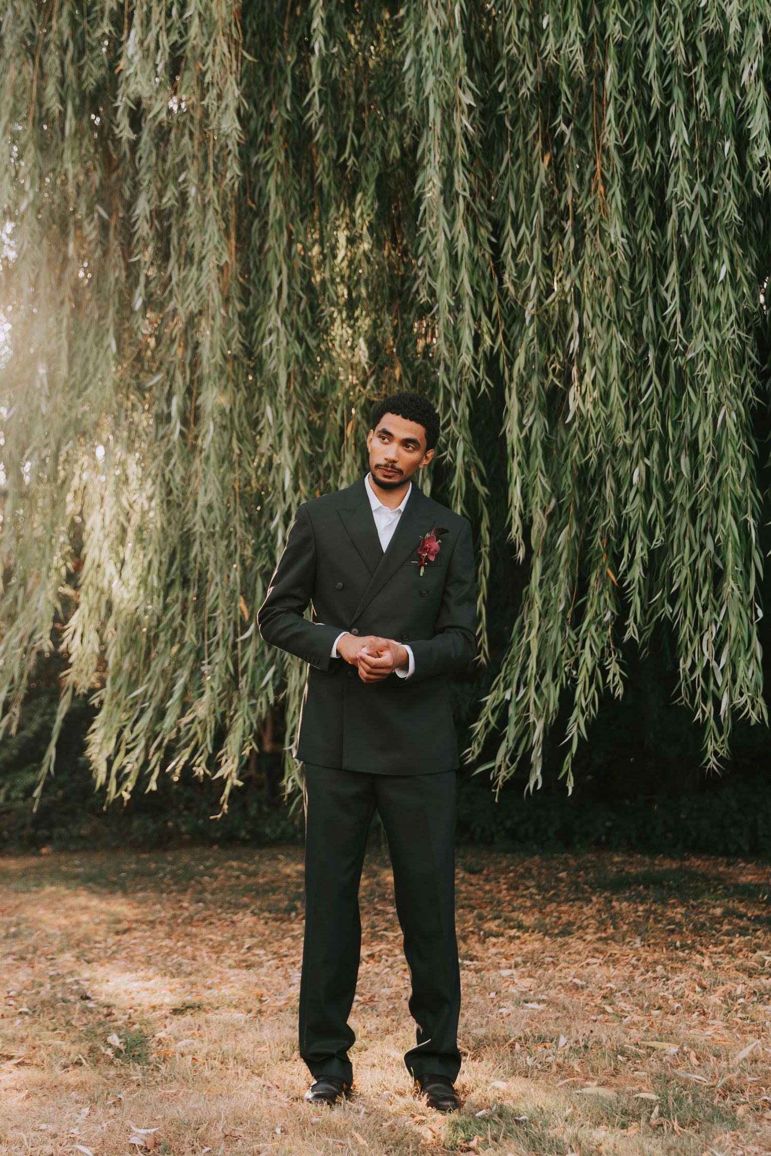 Groom in dark double-breasted suit with boutonniere stands under weeping willow tree outdoors.