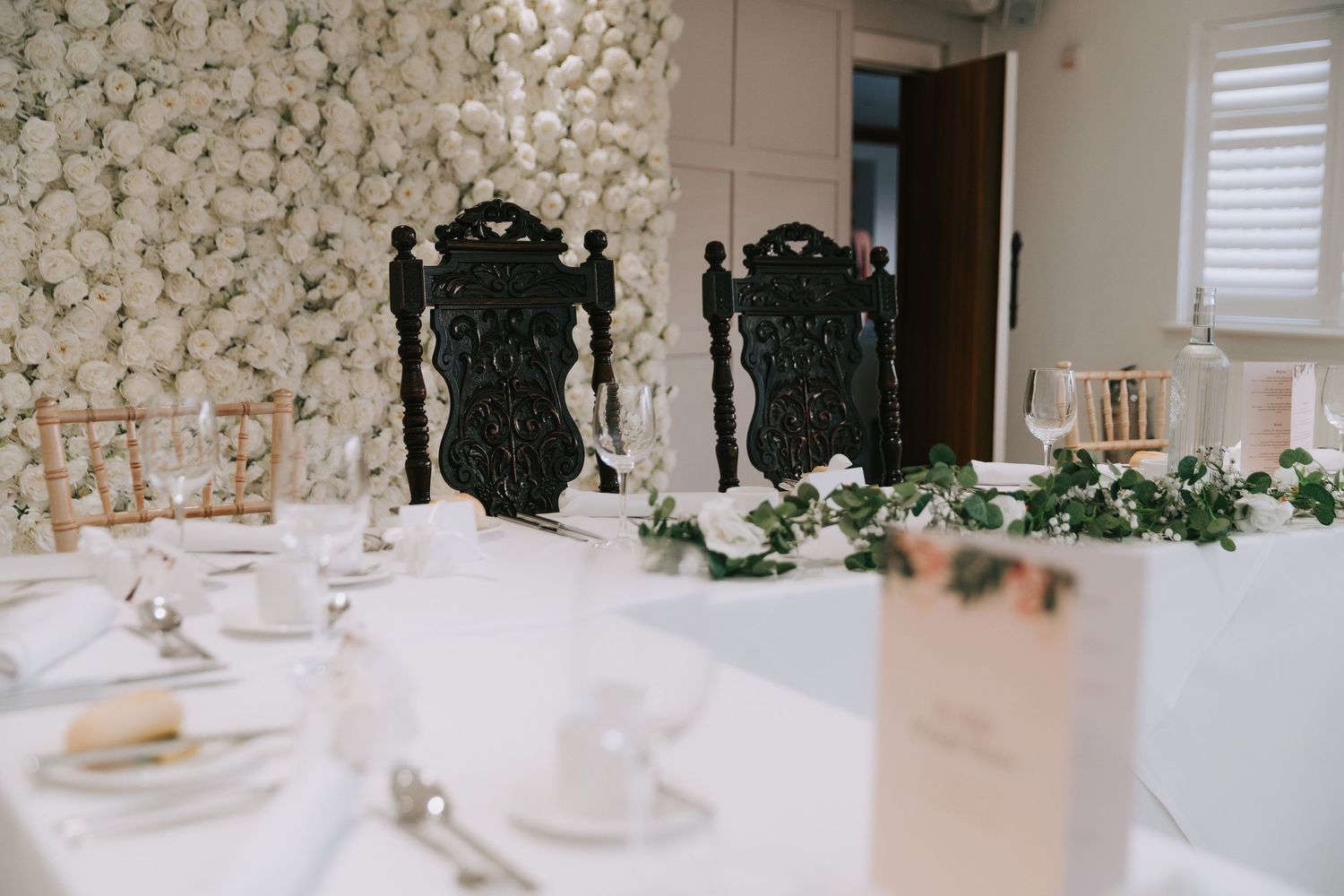 Elegant wedding reception table with white floral wall backdrop, ornate black chairs, and lush greenery centerpiece.