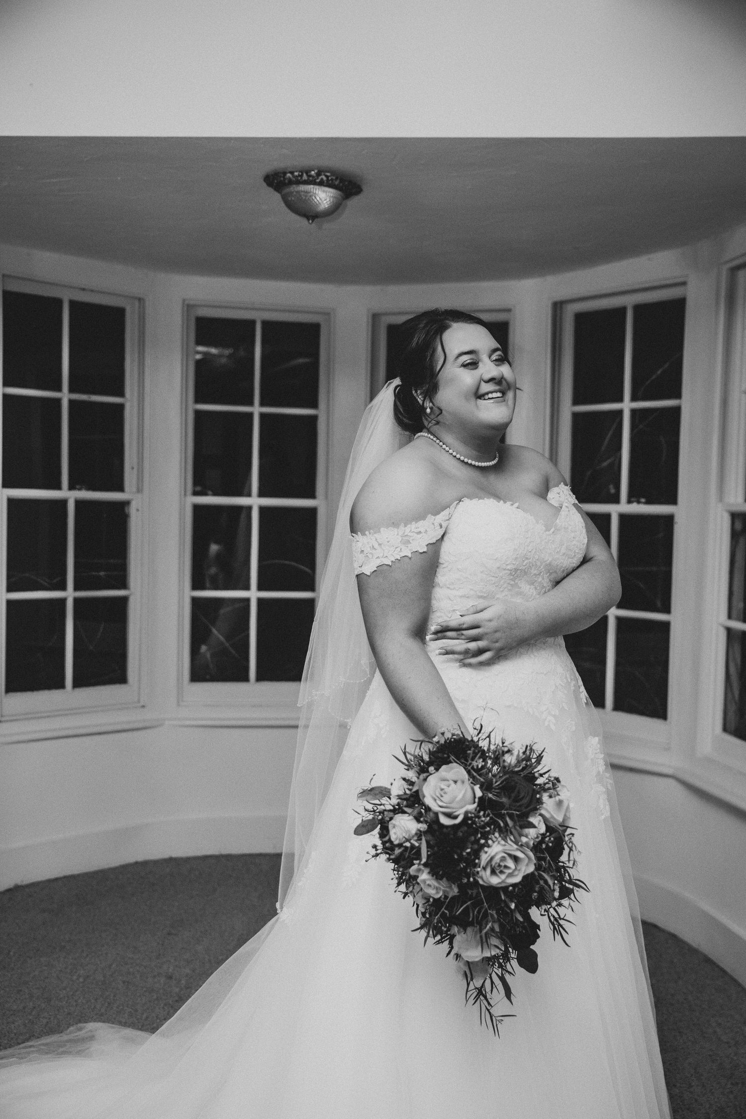 Black and white bridal portrait of a smiling bride in an off-shoulder gown holding a bouquet in a bay window.