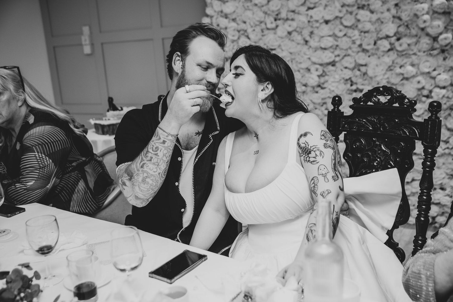 Black and white photo of tattooed bride and groom sharing a sweet moment at their wedding reception table.