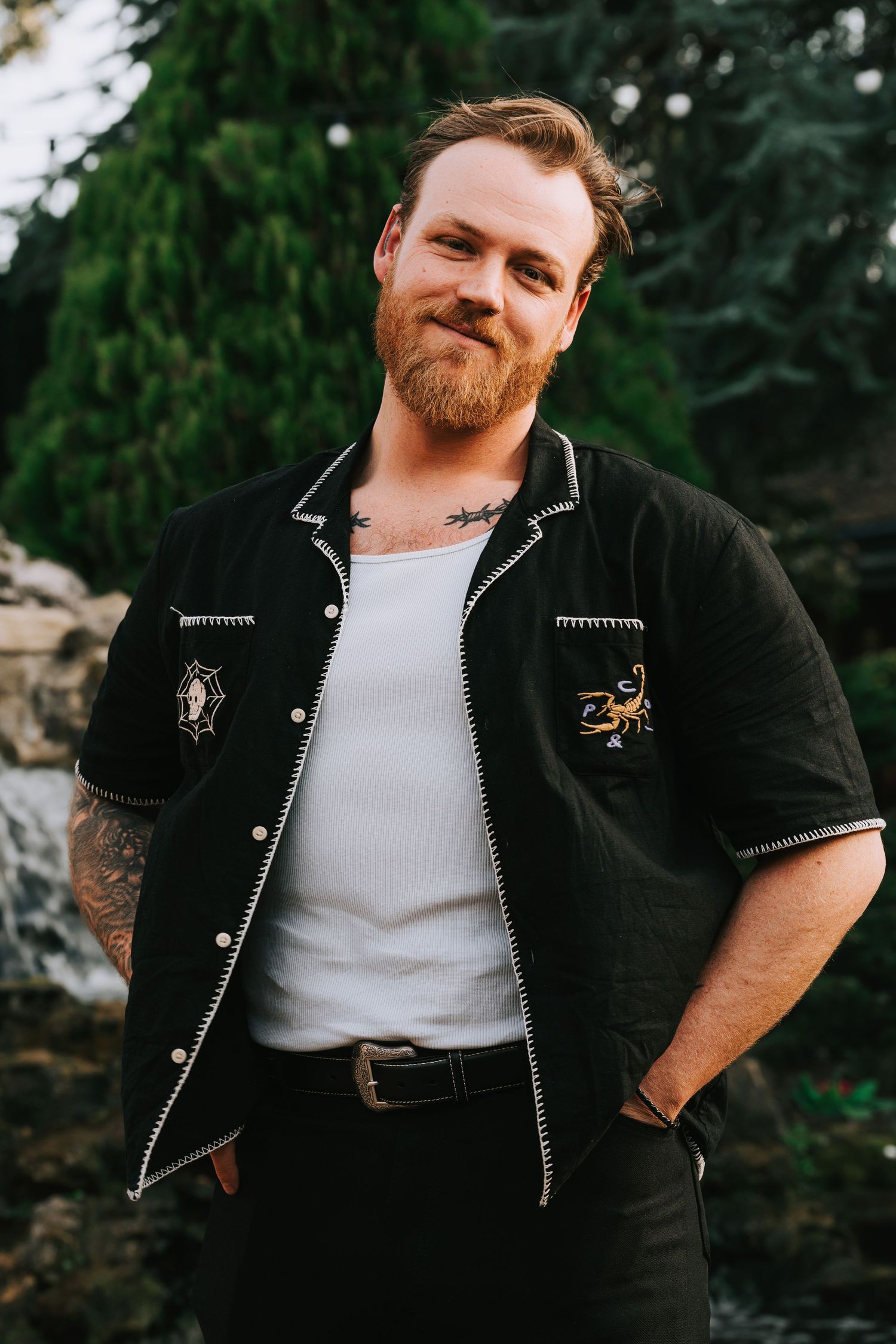 Smiling bearded man with tattoos wearing a black embroidered shirt over white tee, standing outdoors near evergreen trees.