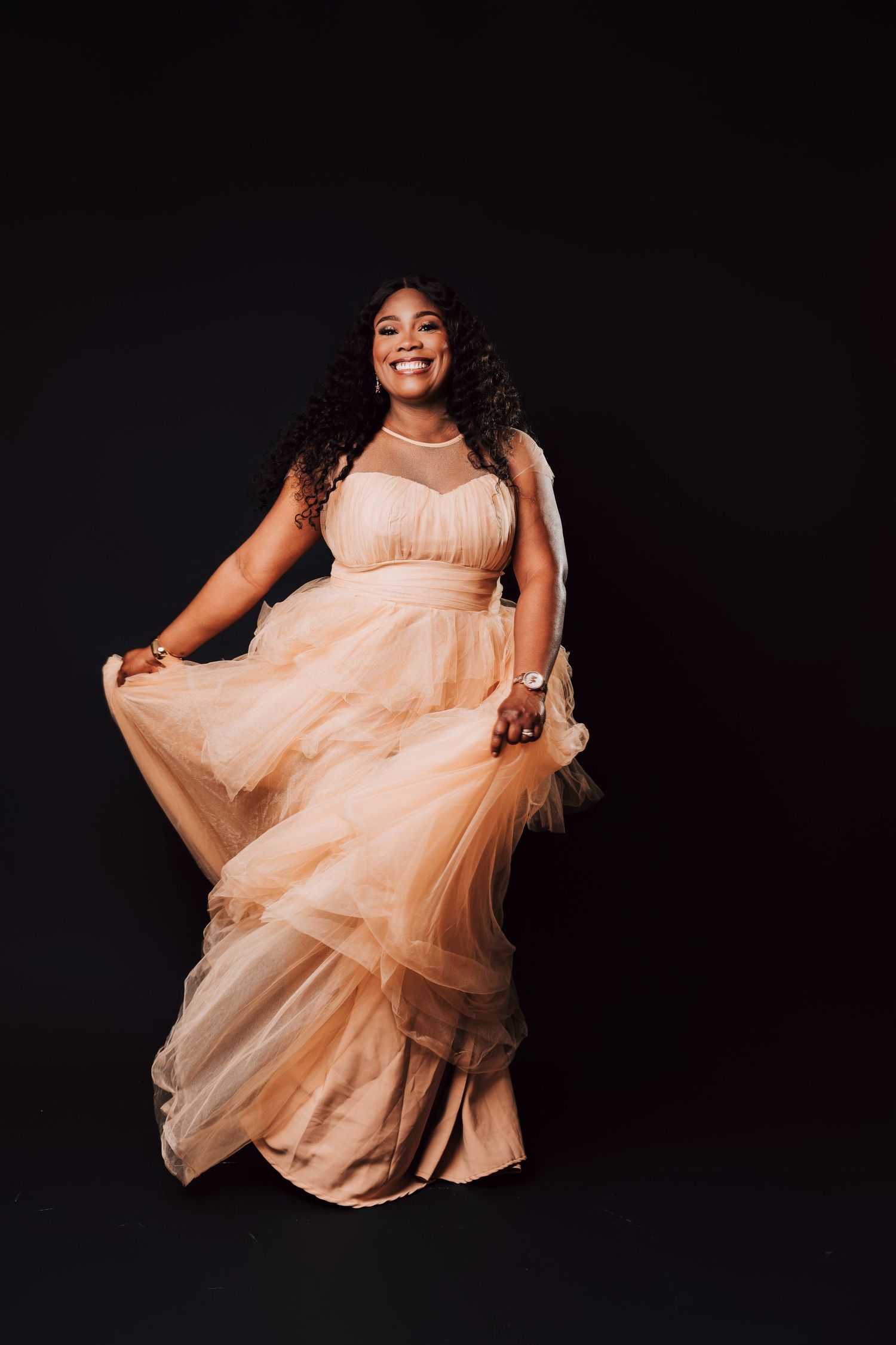 Smiling woman in elegant peach tulle gown posing gracefully against a black studio background.