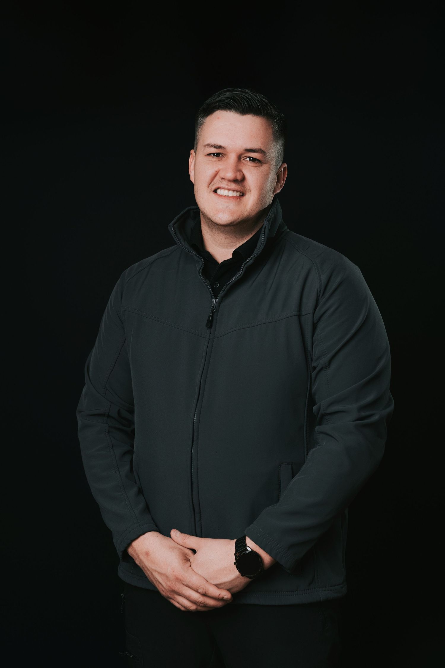 Smiling man in dark green zip-up jacket posing against black background with hands clasped.