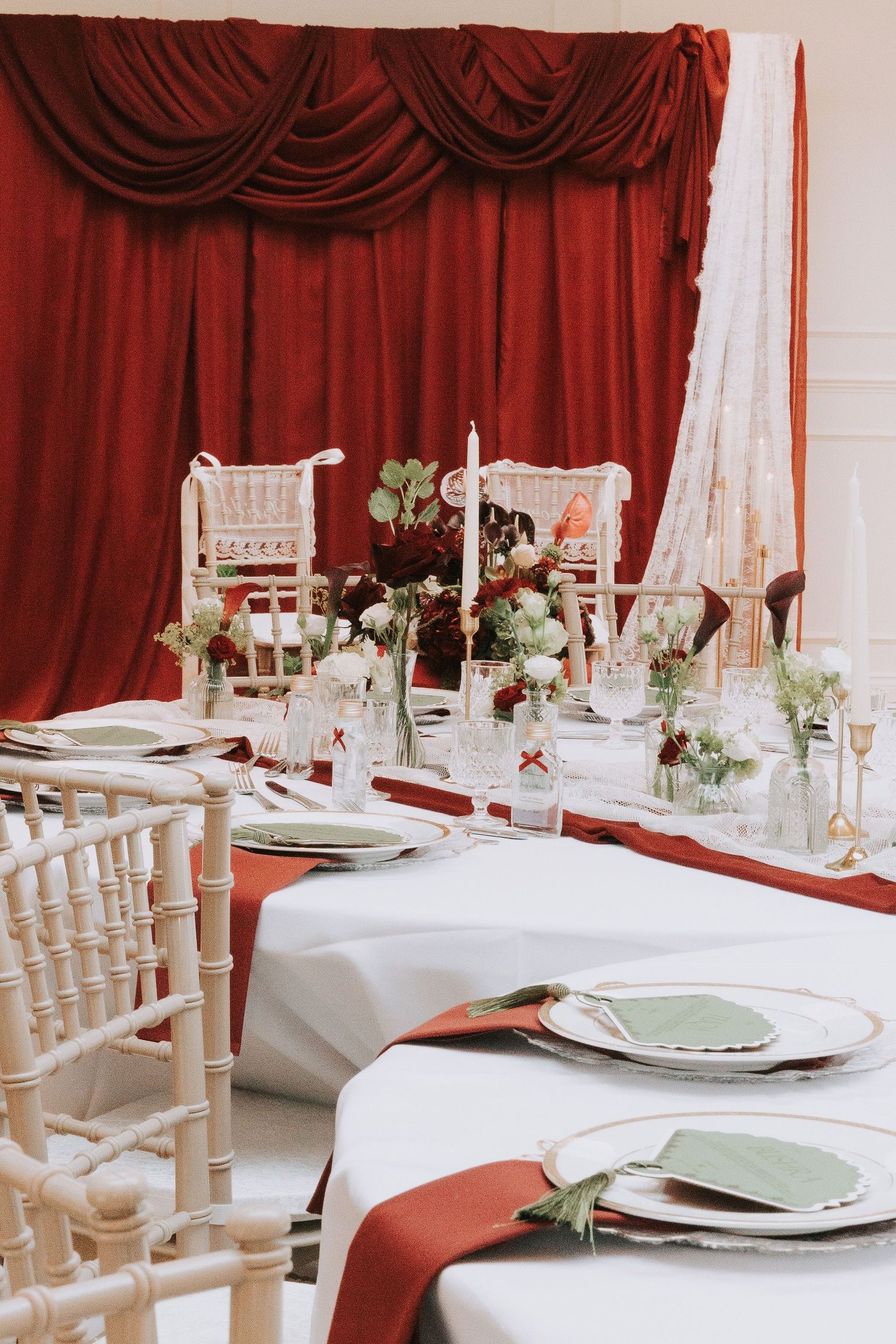 Elegant wedding reception table with white linens, red satin runner, floral centerpieces, and draped burgundy curtain backdrop.