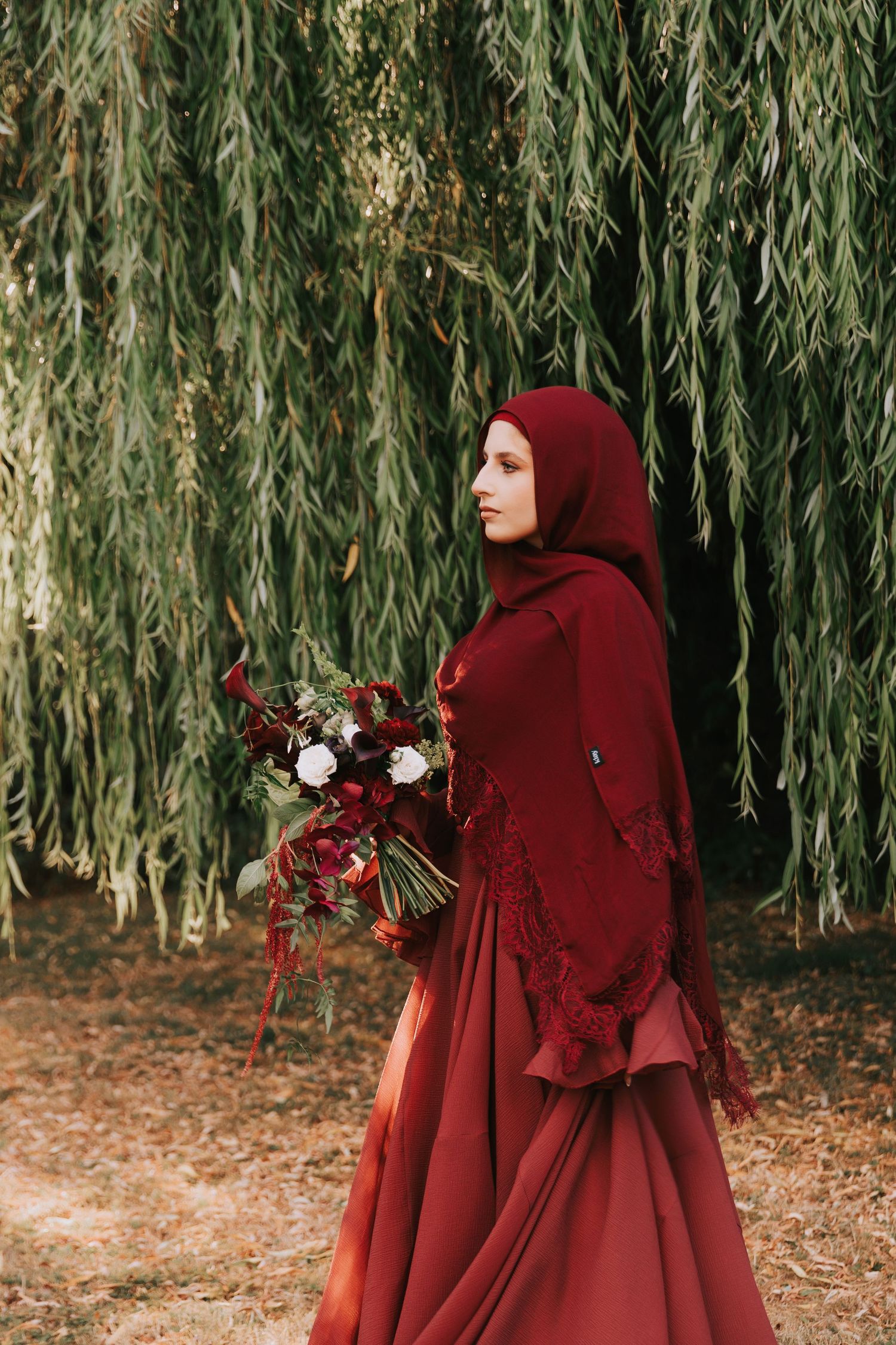 Woman in burgundy hijab and gown holds red and white bouquet under weeping willow tree.