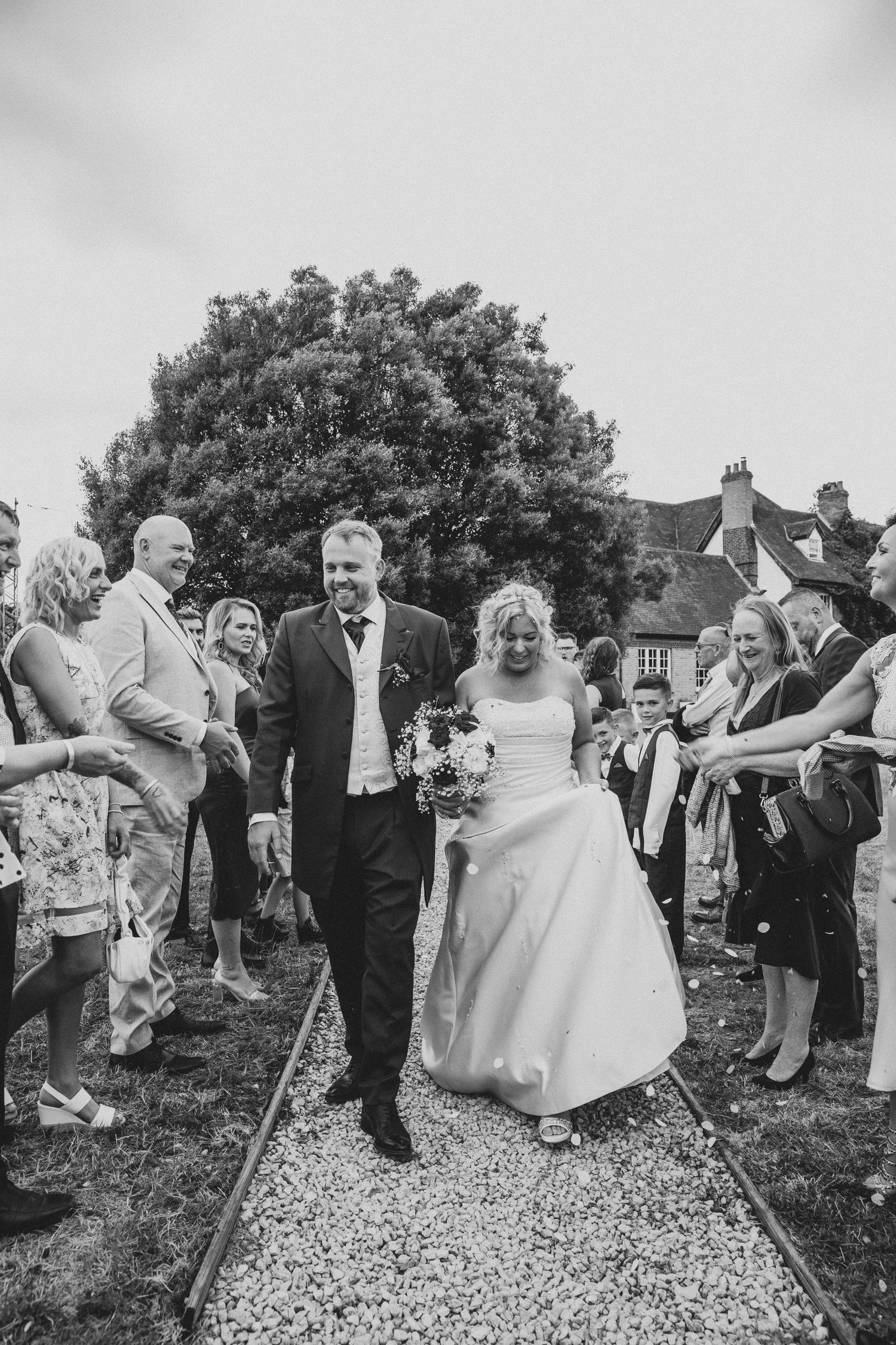 Black and white photo of newlyweds walking through confetti tunnel surrounded by cheering wedding guests outdoors.