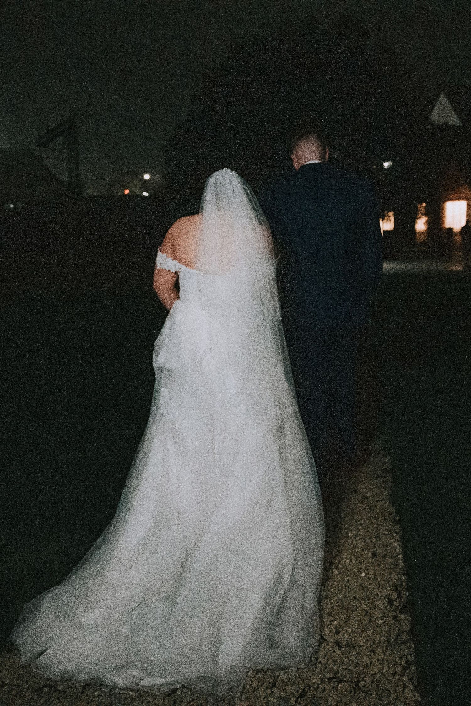 Bride in white gown and veil walks with groom in navy suit at night during outdoor wedding reception.