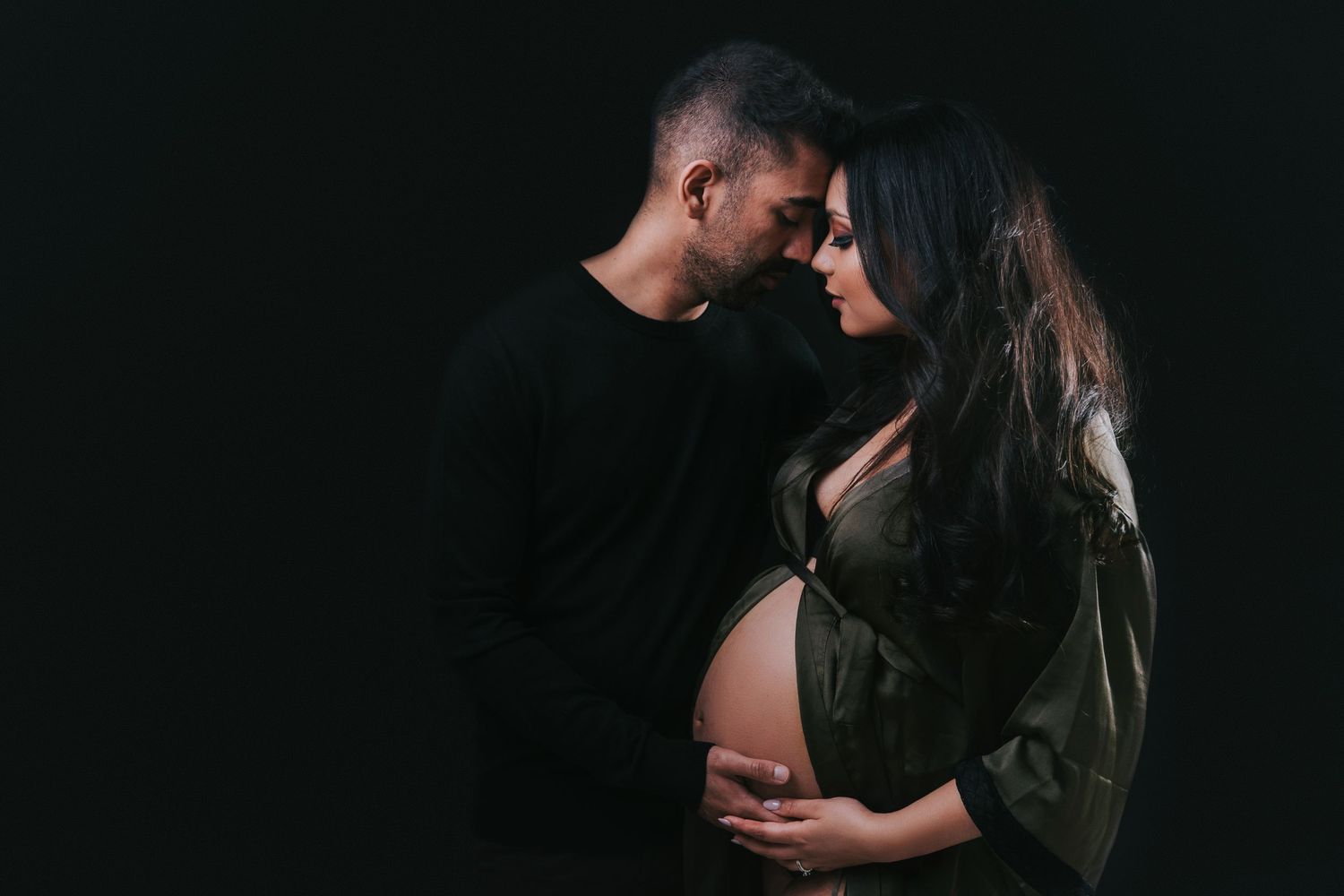A romantic maternity photo of a couple in black clothing against a dark background sharing an intimate moment.