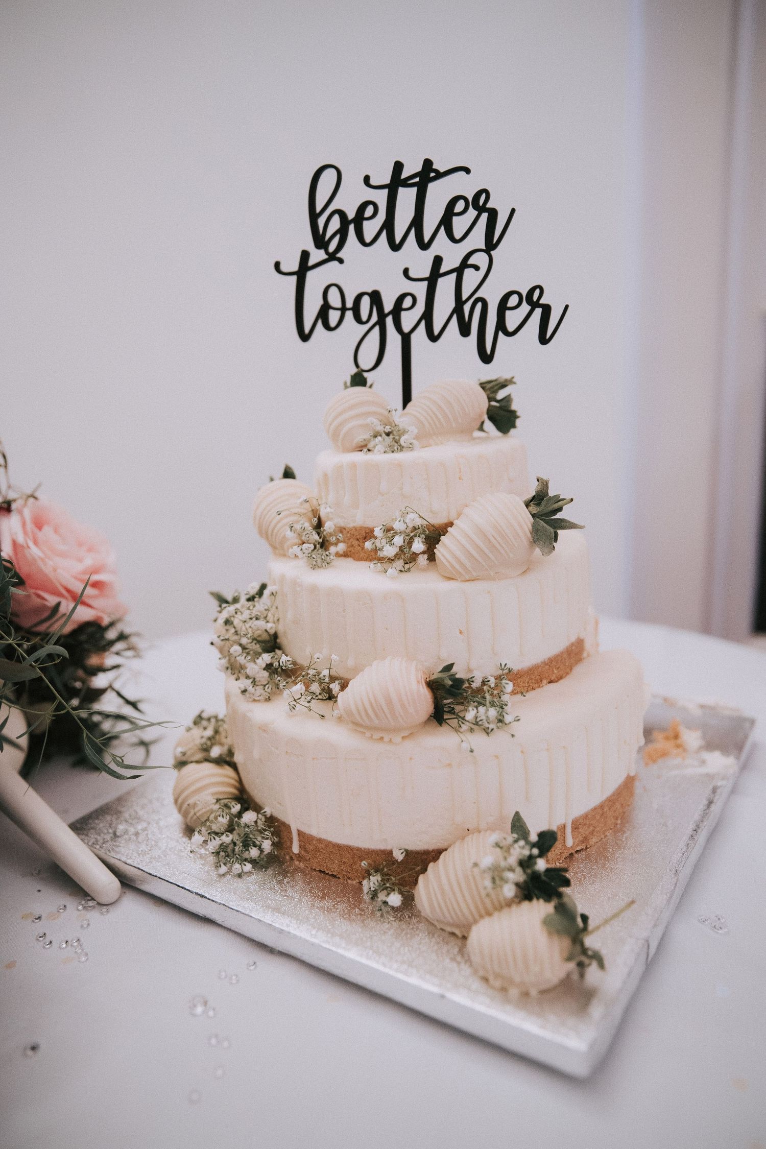 Three-tier white wedding cake decorated with white chocolate strawberries and topped with better together cake topper.