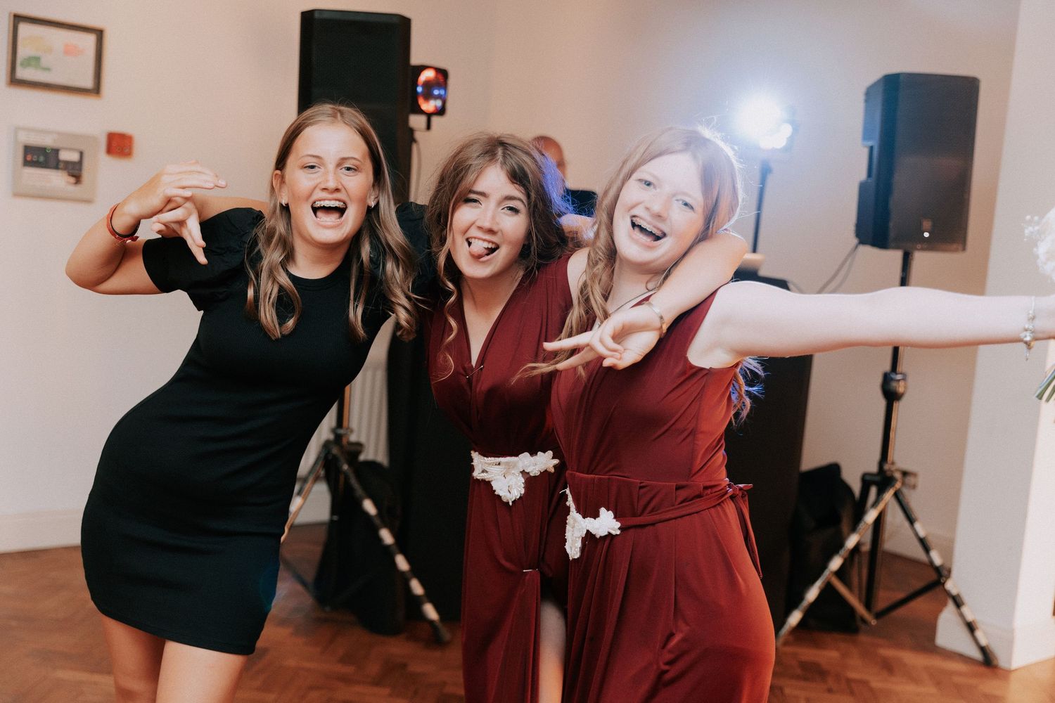 Three women laughing and posing playfully at a party, one in black dress, one in burgundy jumpsuit, one in white outfit.