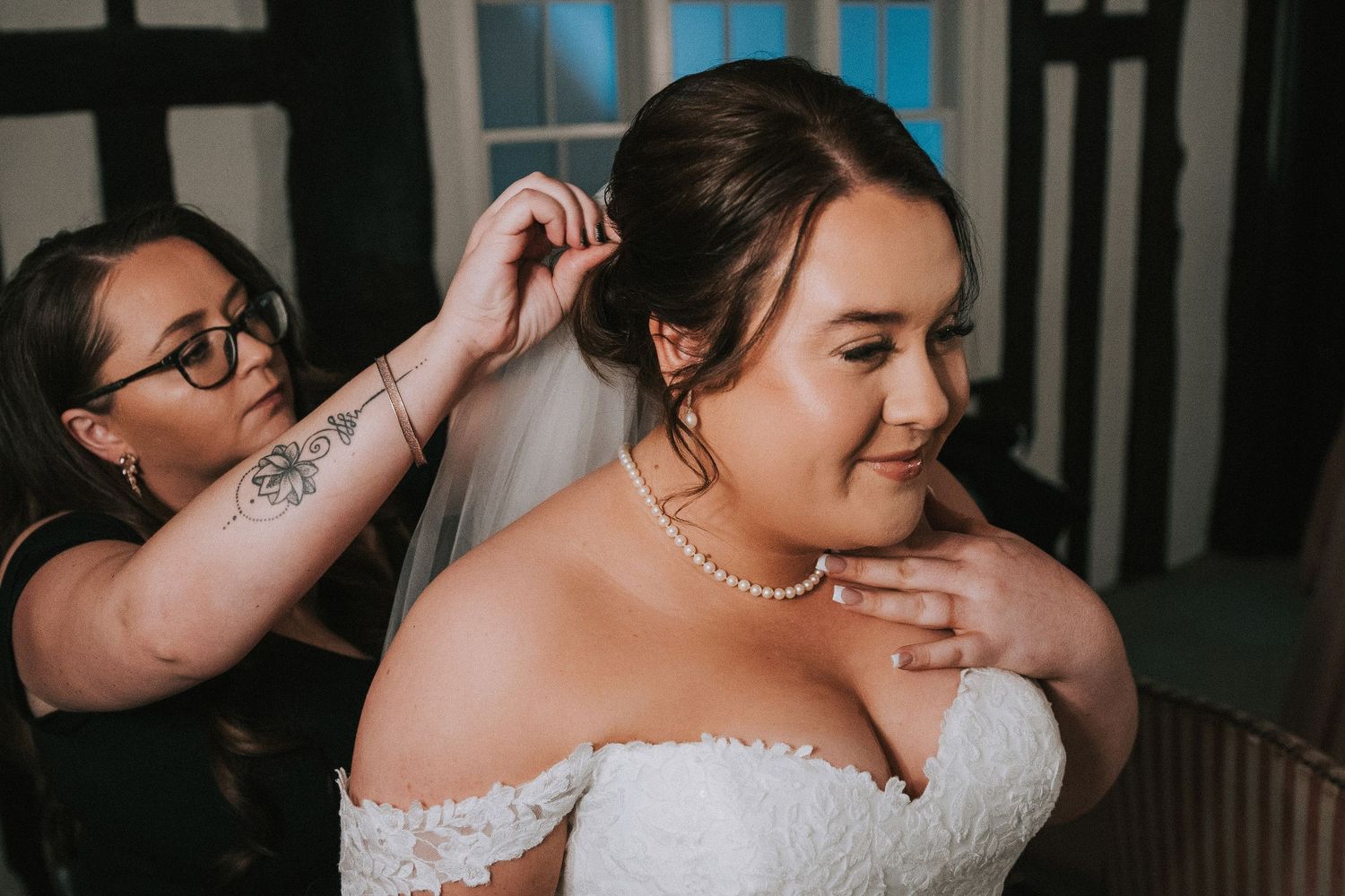 Hair stylist with tattoos attaching a veil to bride in off-shoulder lace wedding dress with pearl necklace.
