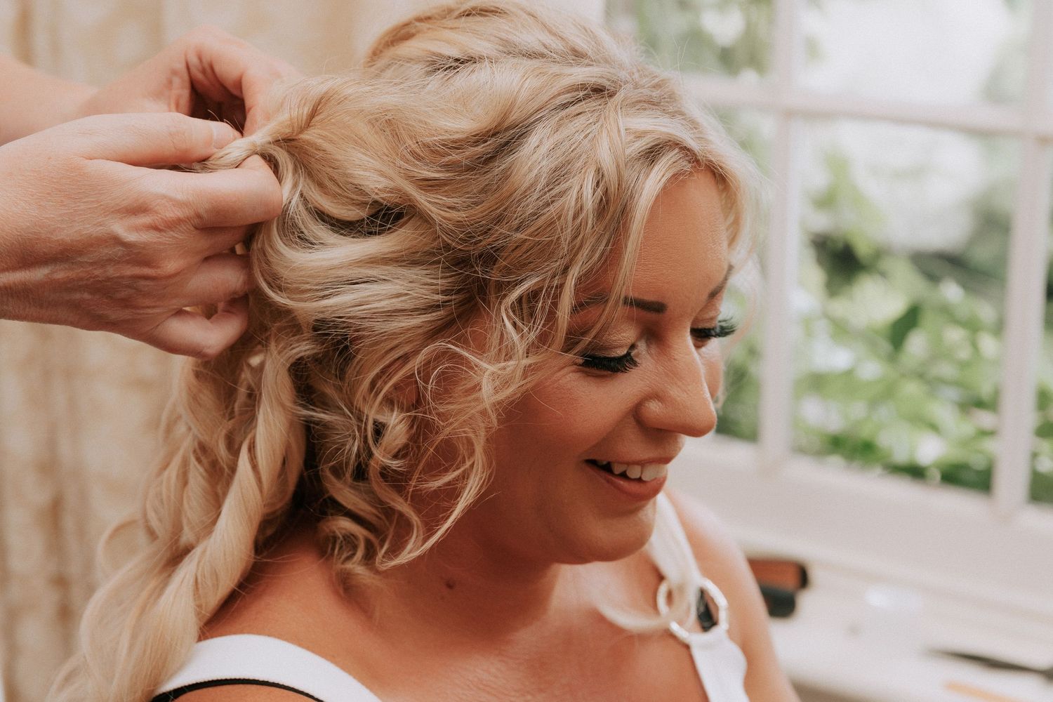 A smiling blonde woman getting her curly updo styled by a hairstylist at a bright window.