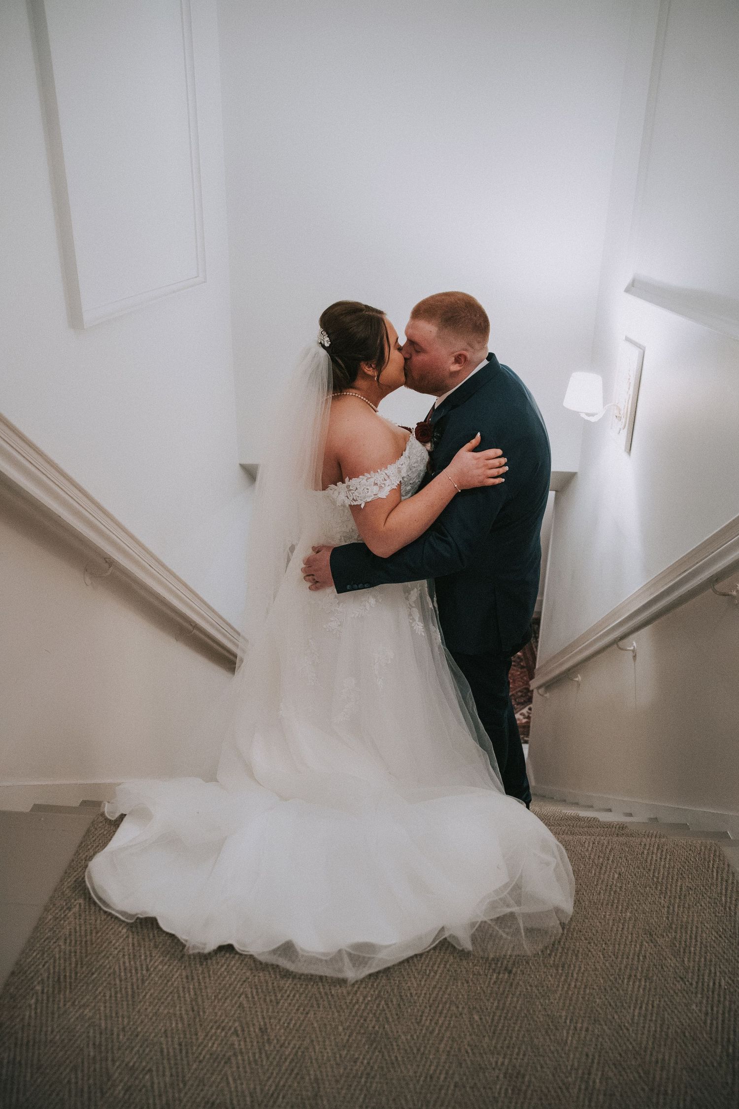 Bride and groom share romantic kiss on staircase, bride in off-shoulder white gown with flowing veil.