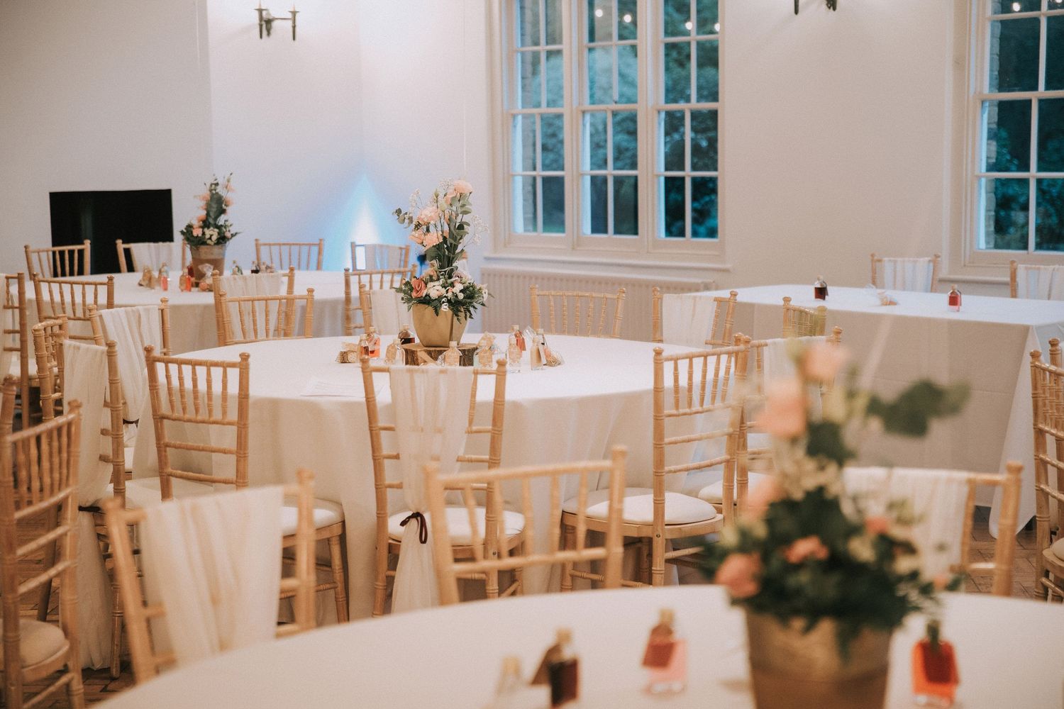 Elegant wedding reception hall with round tables, white linens, chiavari chairs, and floral centerpieces in a bright venue.