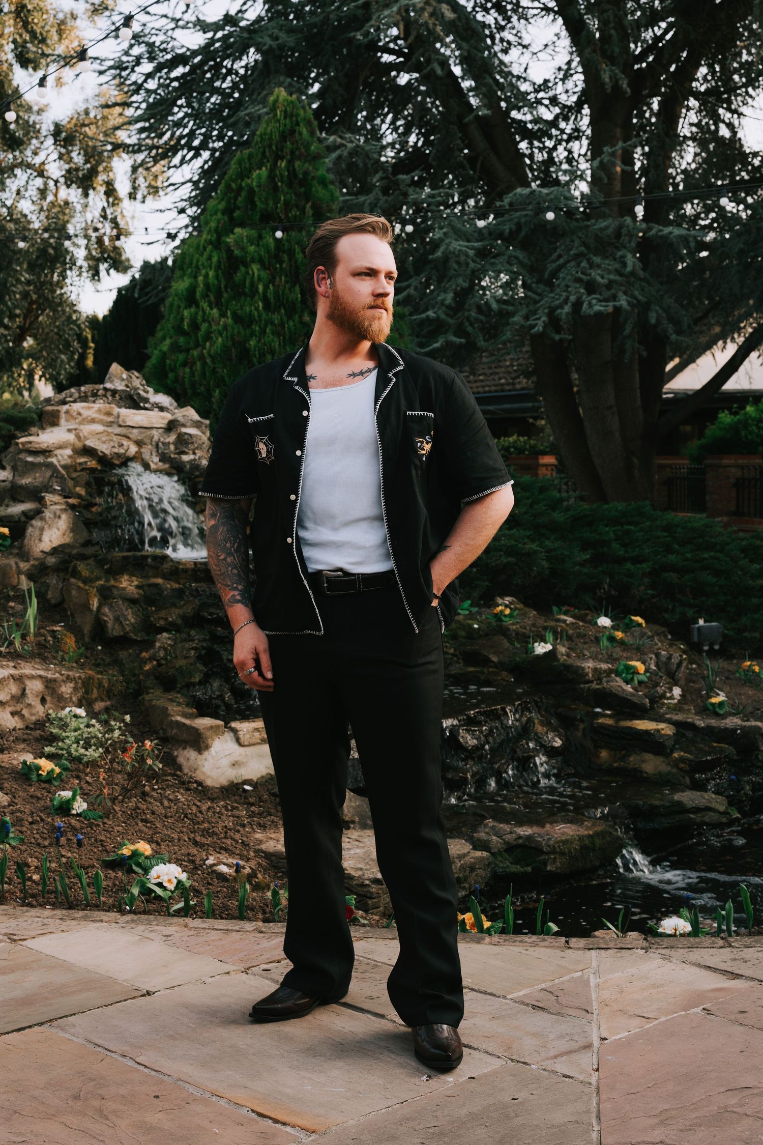 A bearded man in a black bowling shirt and dark pants stands confidently at Ye Olde Plough House  gardens with stone waterfall backdrop.