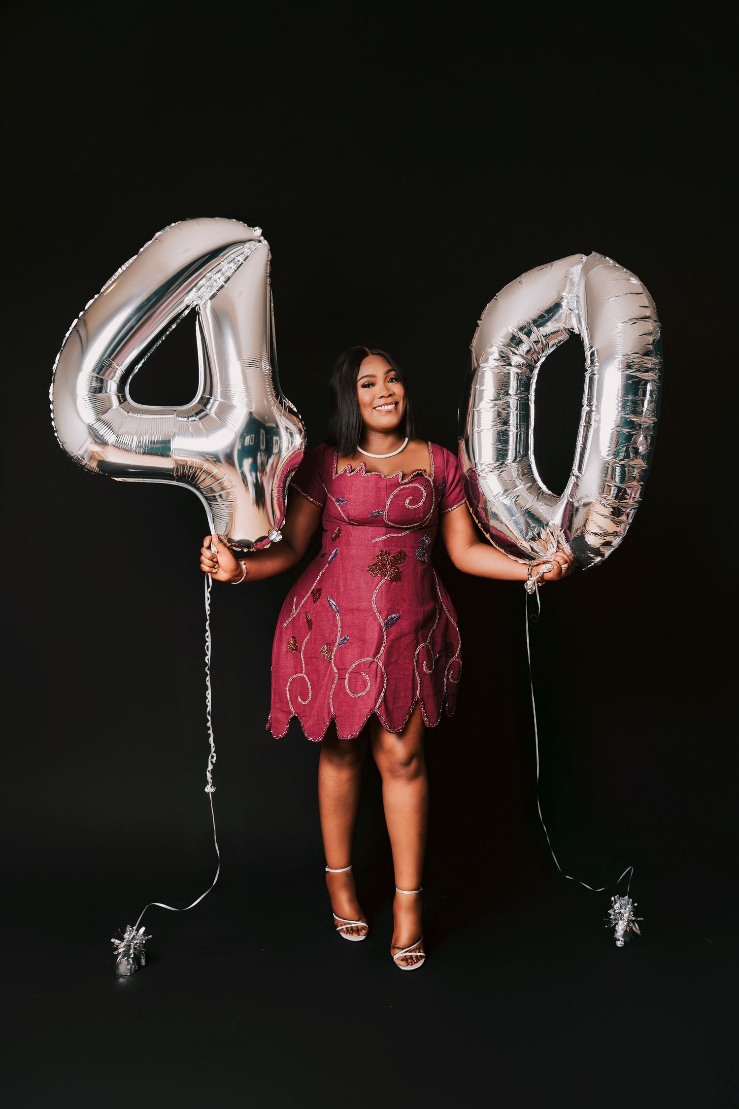 A smiling woman in a pink dress holds large silver number 40 balloons celebrating her 40th birthday against a black background.