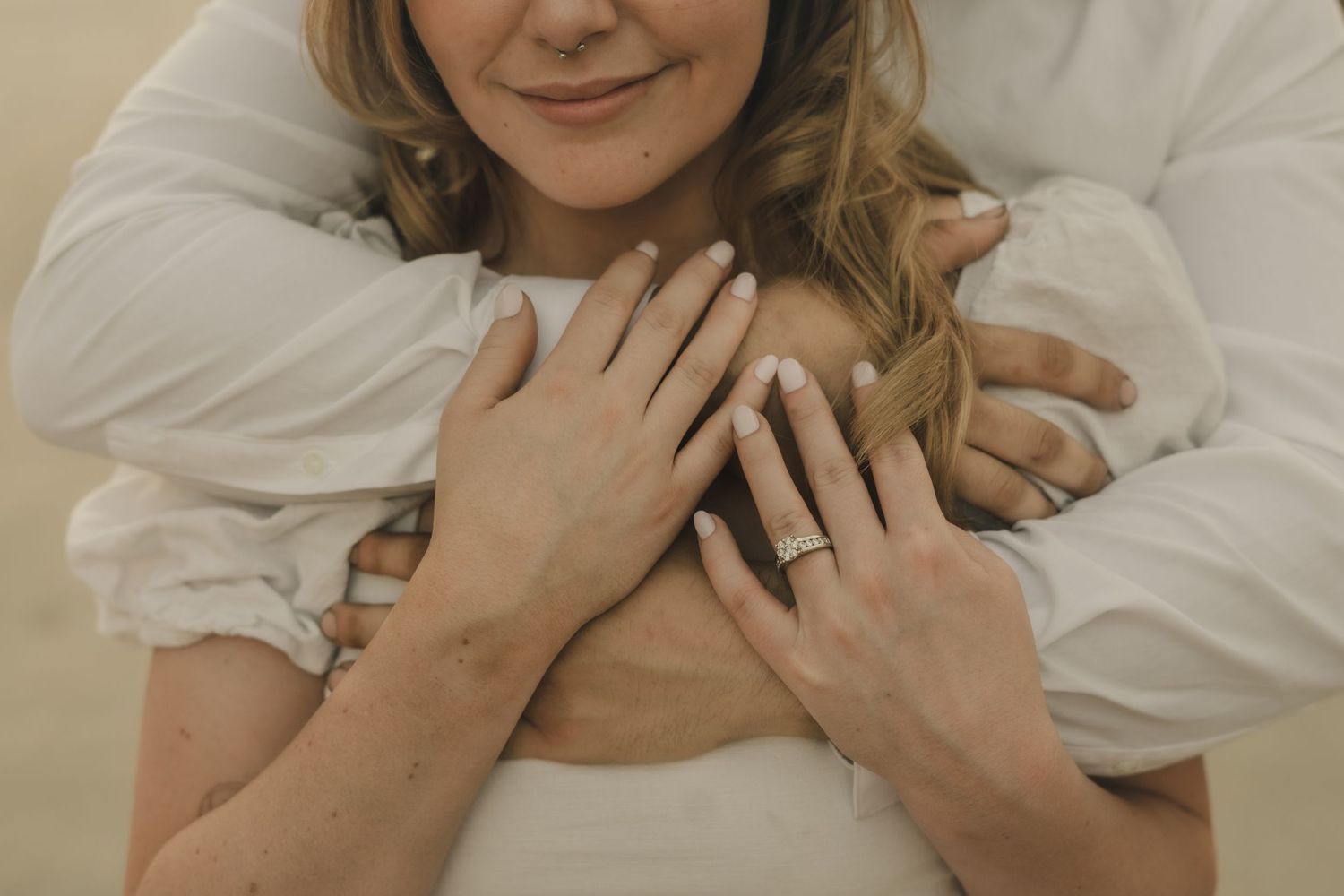 a close up of a bride smiling while her groom wraps his arms around her chest and she holds onto his hands