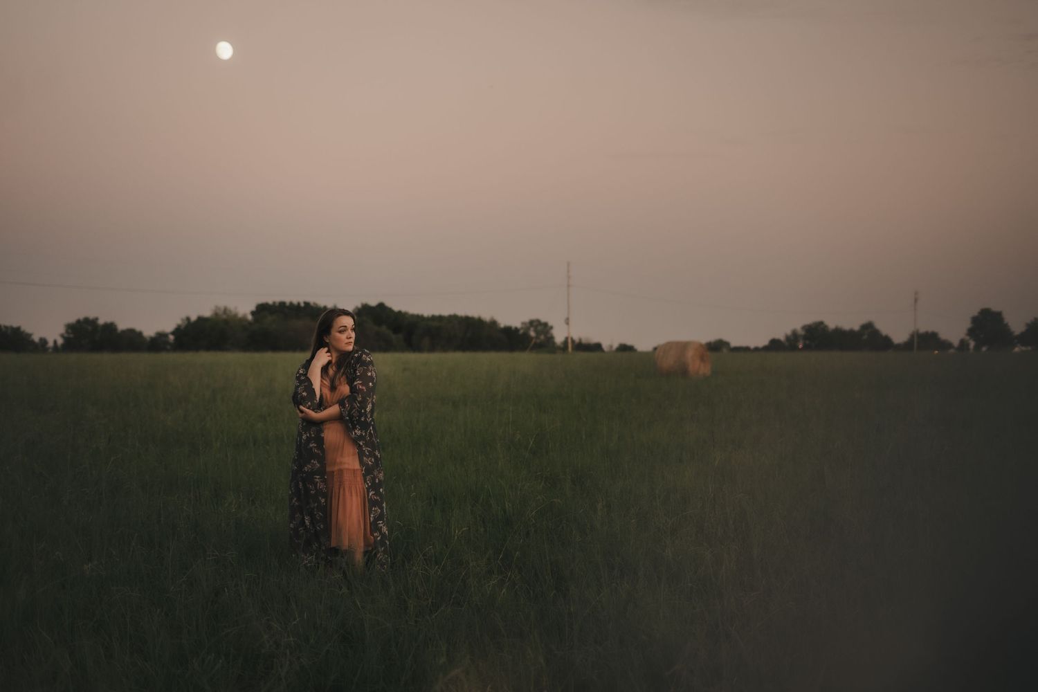 a woman in a floral dress standing in a grassy field with the moon in the sky
