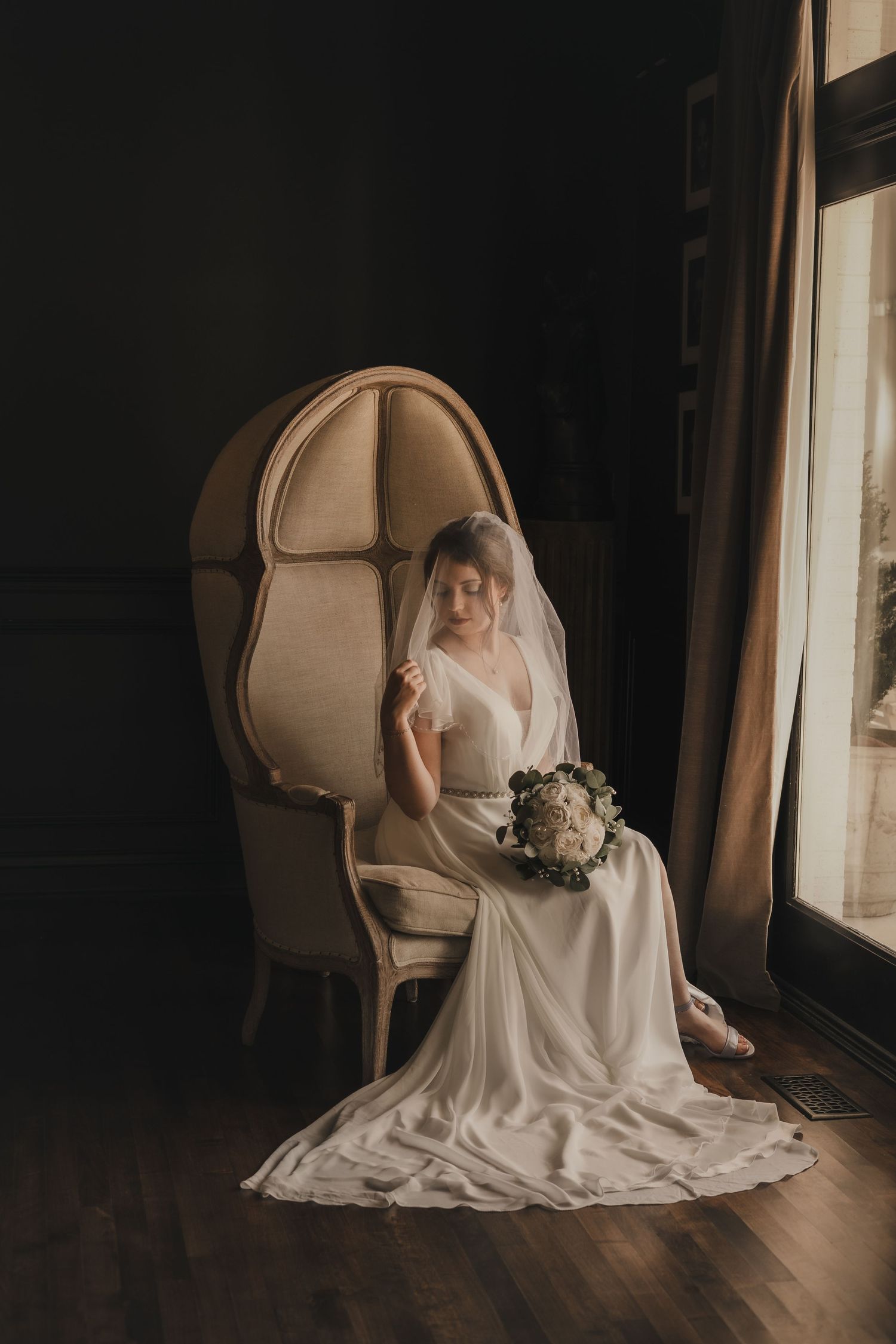 a bride sitting in an elegant chair in front of a window while holding a bouquet and looking down