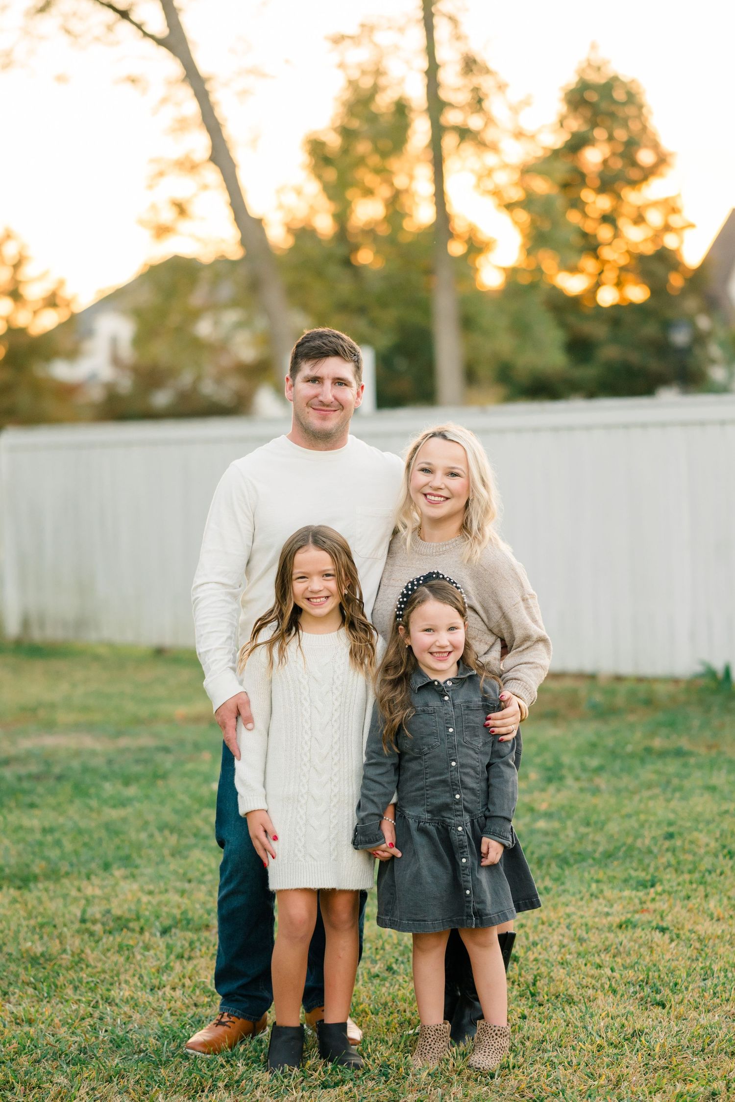 A family in coordinated cream and gray outfits poses together on grass during golden hour sunset.
