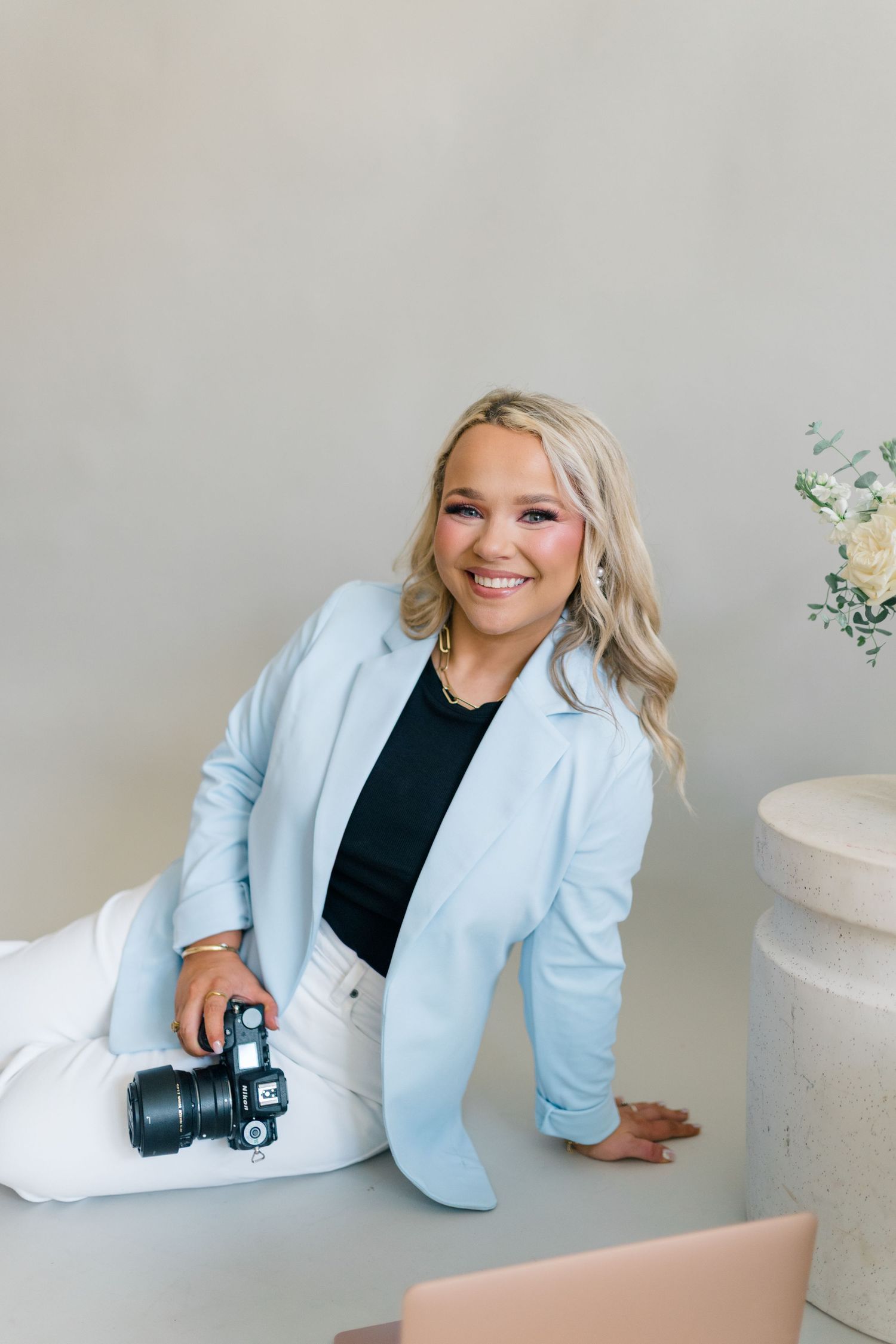 Smiling blonde woman in light blue blazer holding a DSLR camera, seated in a bright studio setting.