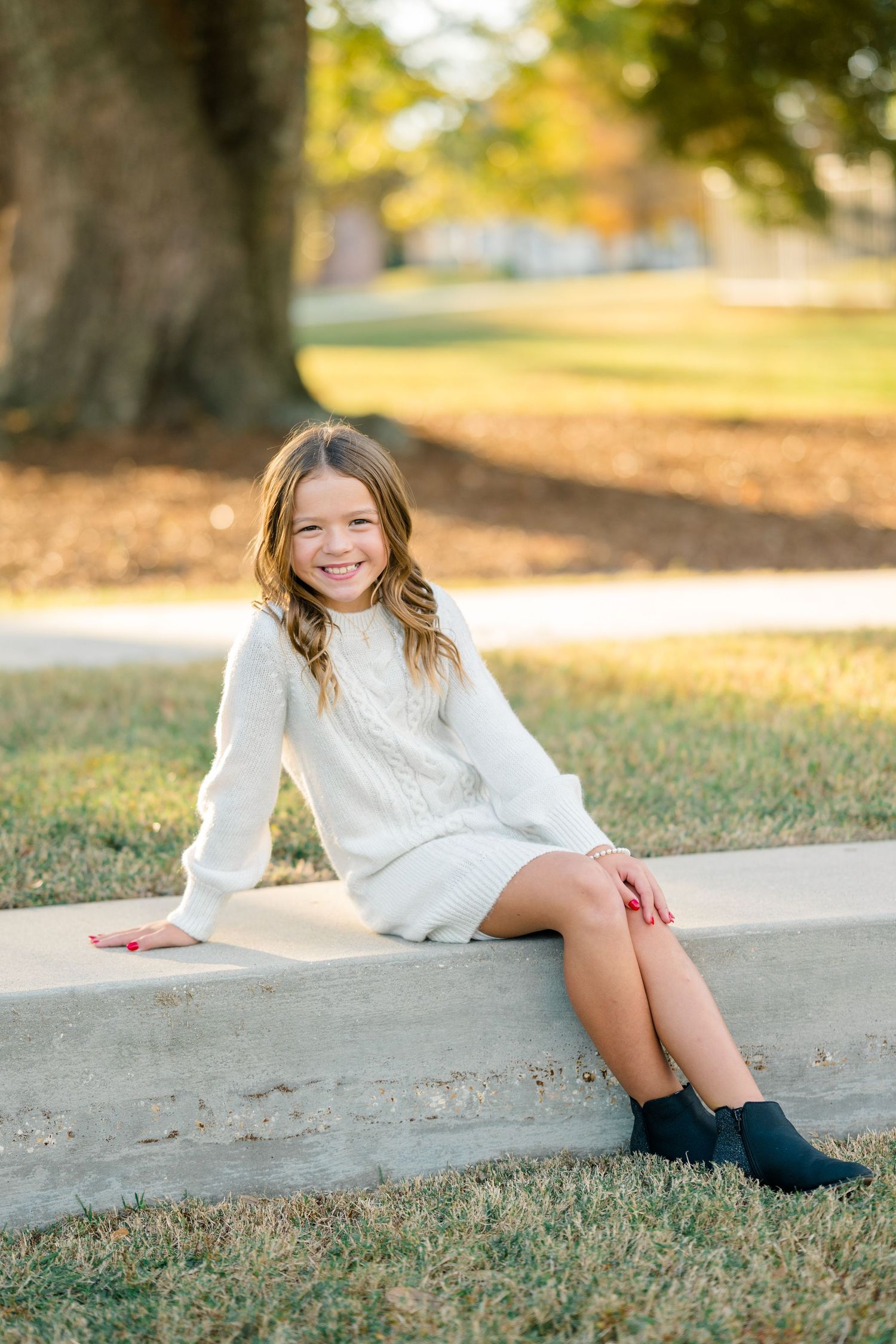 A person in a white sweater poses on grass near a curb during a sunny autumn day with golden leaves in the background.