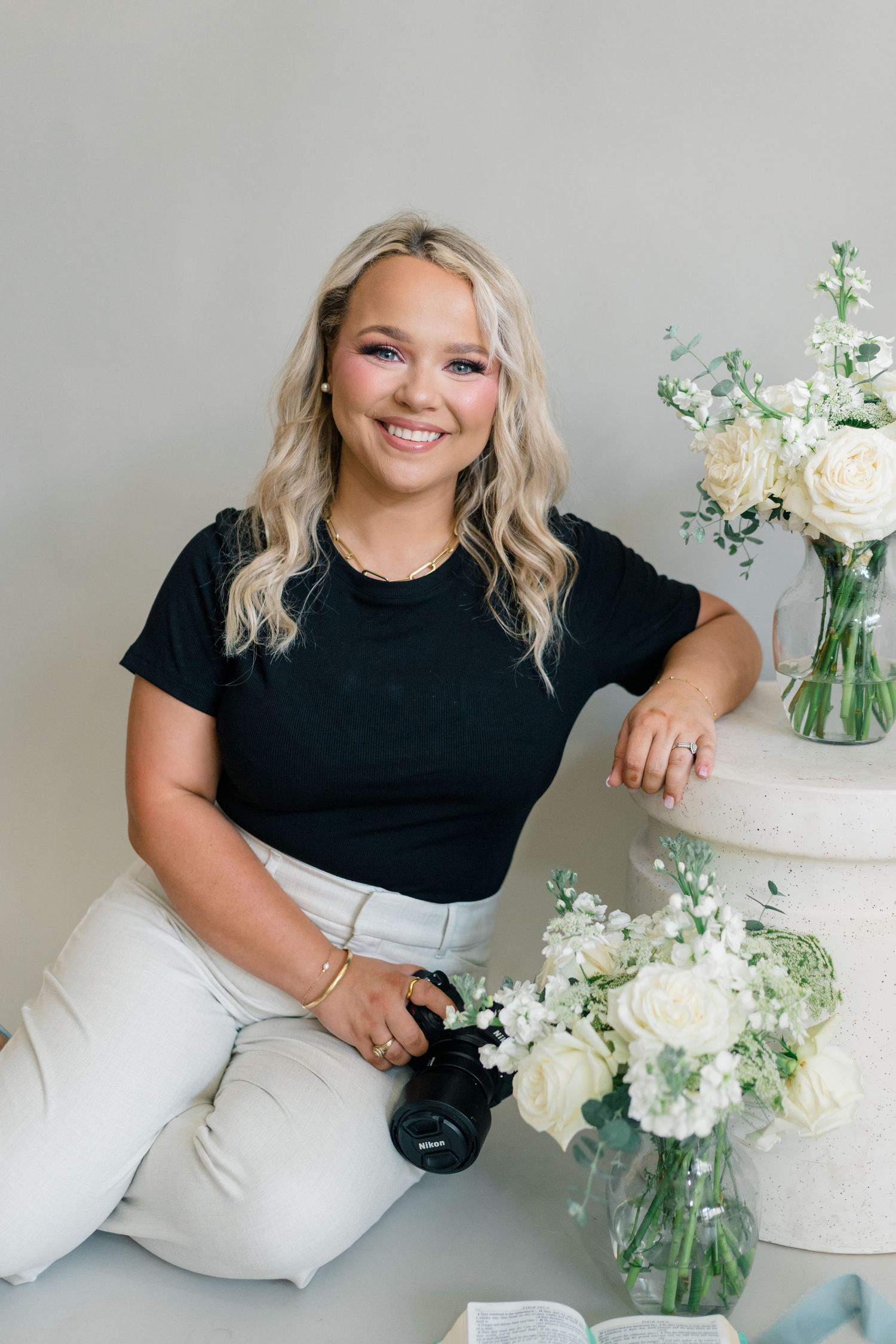 Smiling blonde woman in black top holding camera seated beside white rose floral arrangements in glass vases.