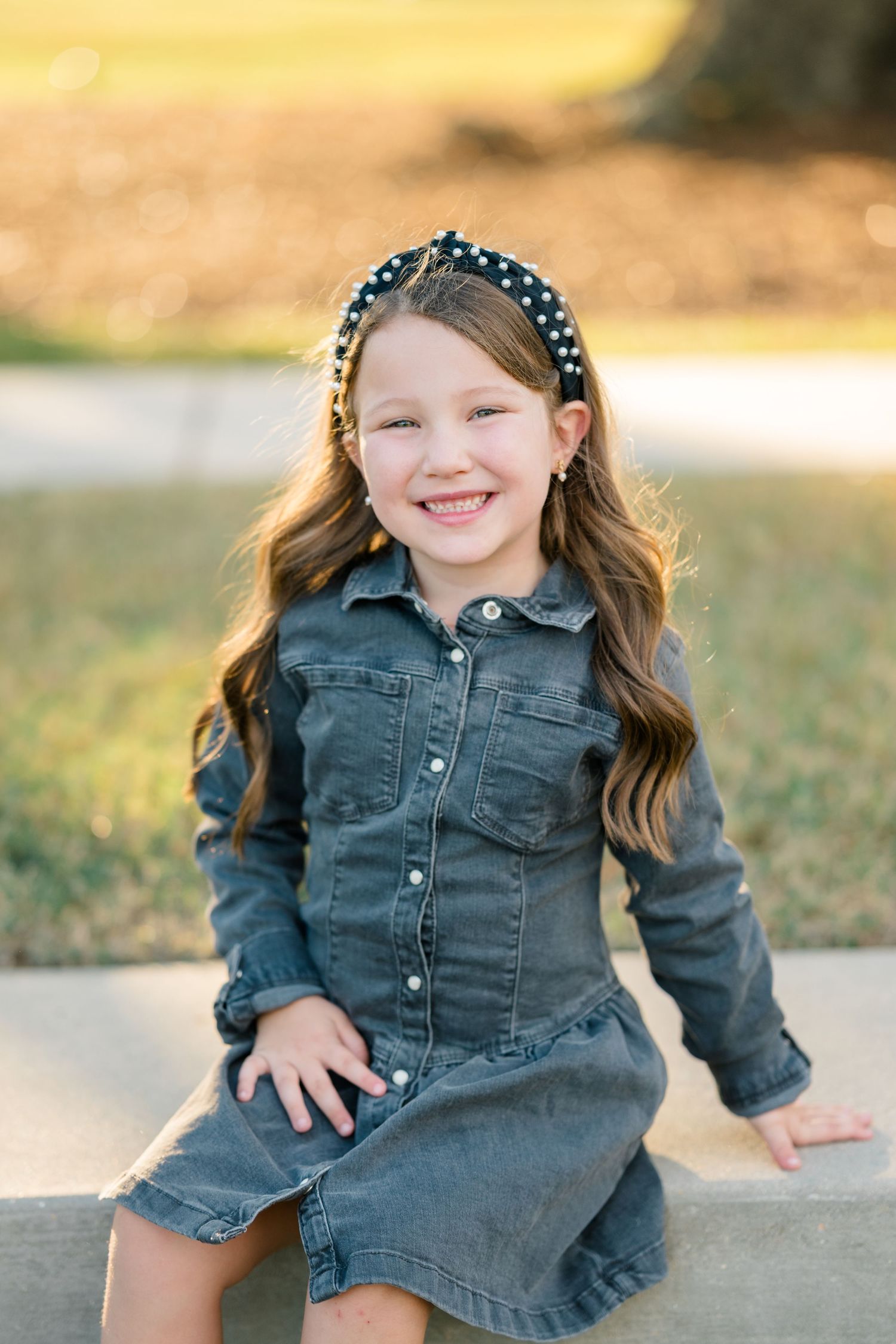 Child wearing denim dress and polka dot headband sits outdoors in warm sunlight.