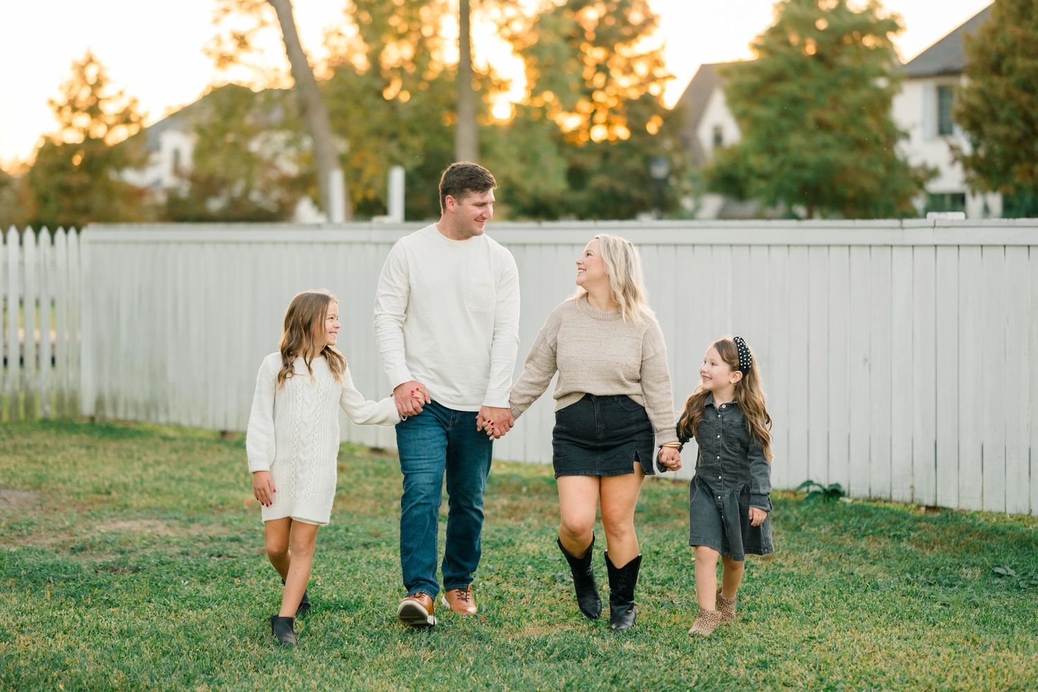 Family walking together on grass in front of white picket fence during golden sunset hour.