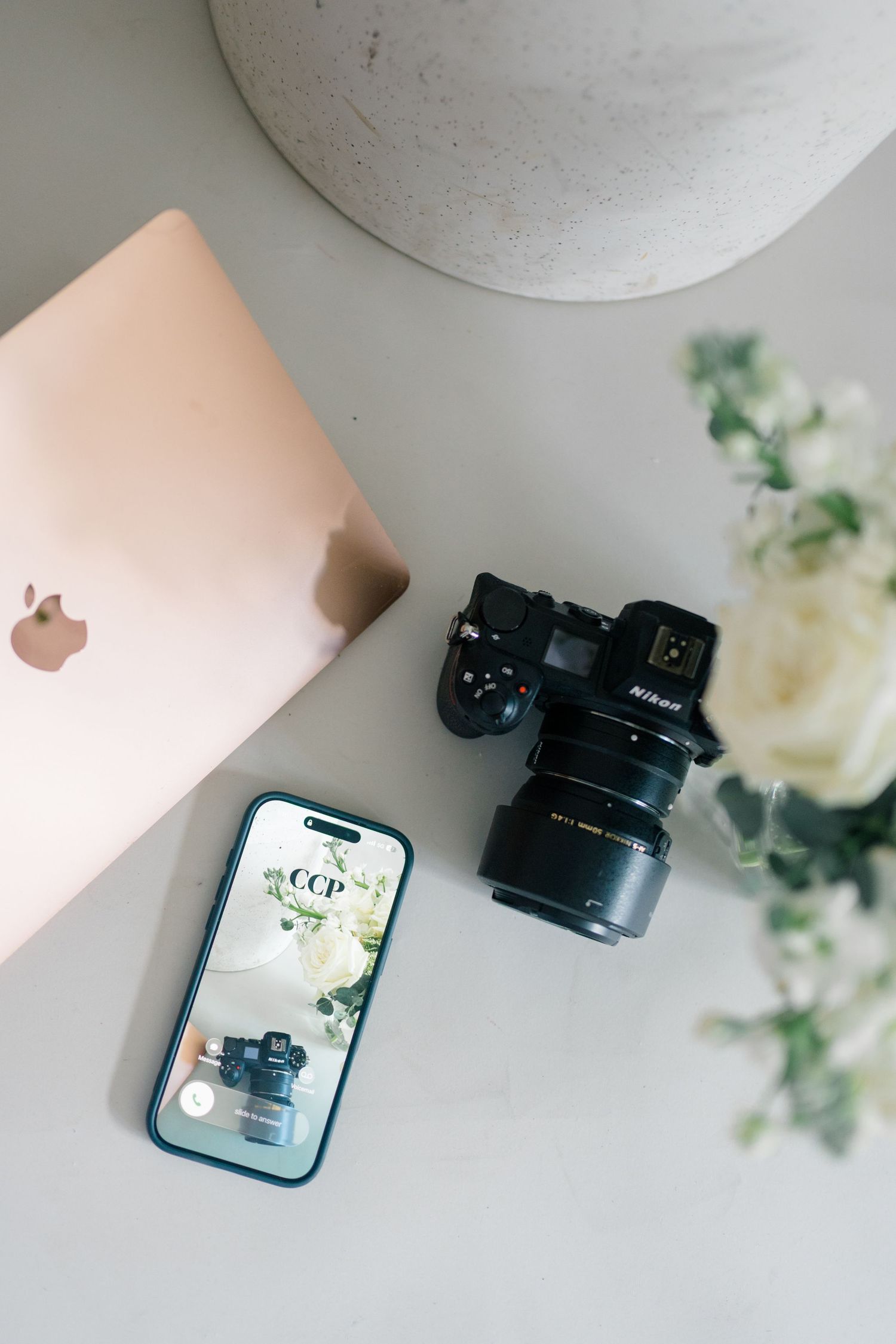 Flat lay of a Nikon camera, iPhone, rose gold MacBook, and white flowers on a gray surface.