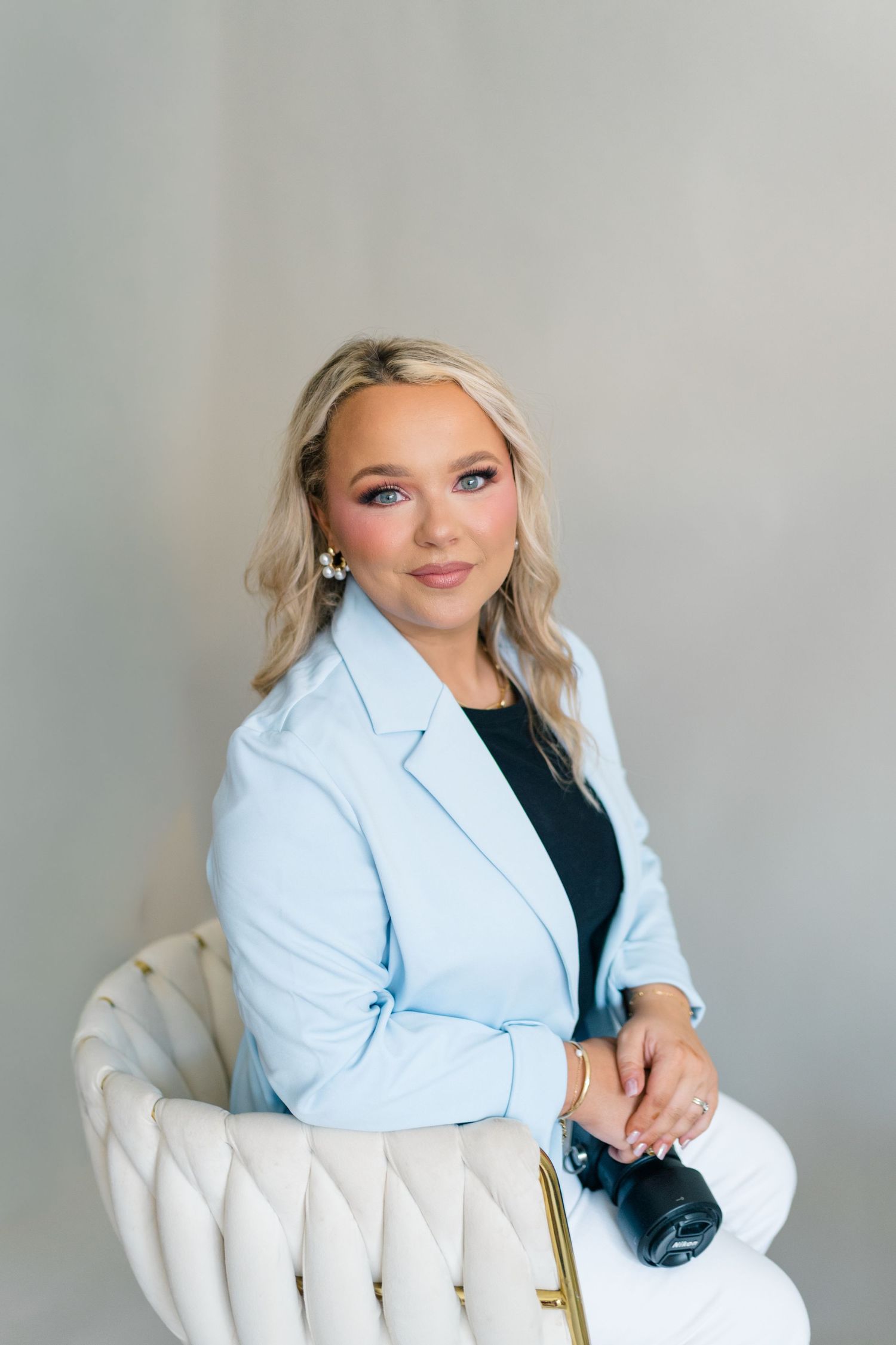 Woman in light blue blazer sits in white tufted chair holding camera, professional portrait photography.