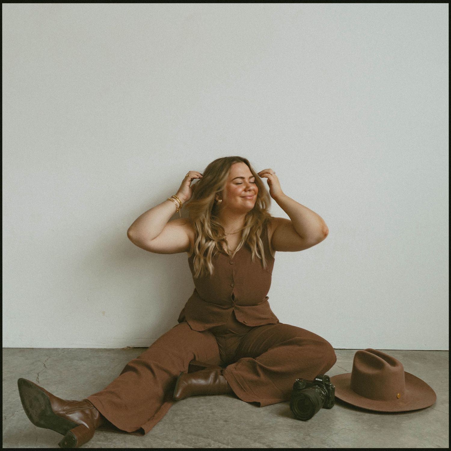 A person in brown clothing sits on the floor against a white wall with a wide-brimmed hat nearby.