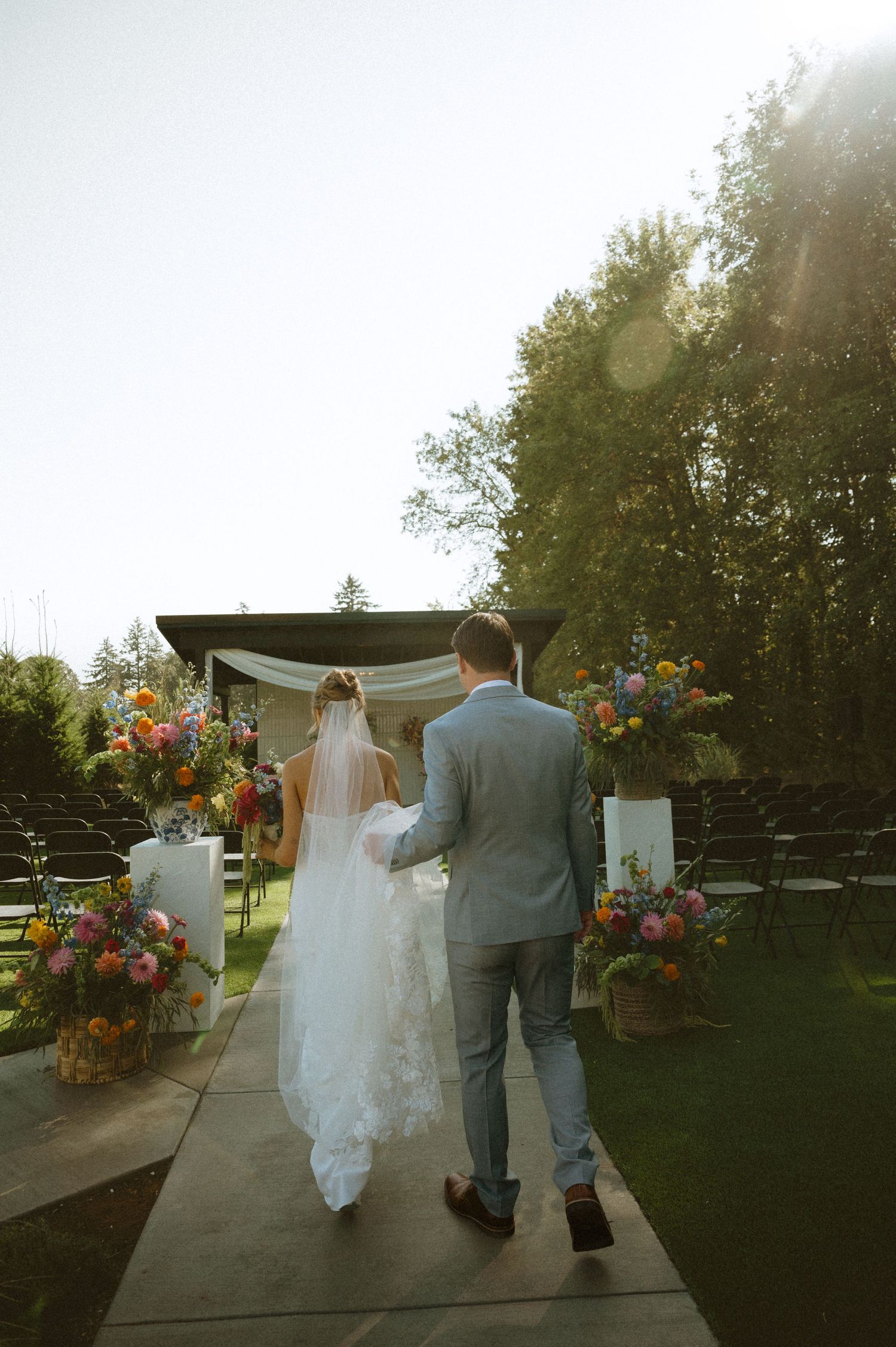 Newlyweds walking down garden path with vibrant flower arrangements in golden sunlight.