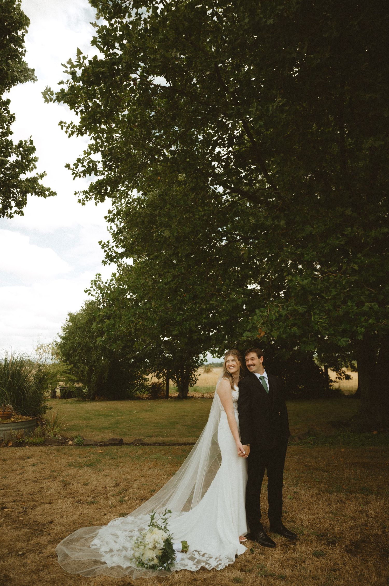Wedding couple standing under large tree in rustic outdoor setting.