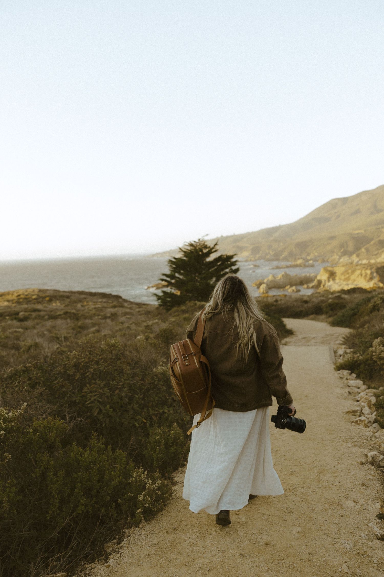 Person in white dress walking along a scenic coastal trail during golden hour.