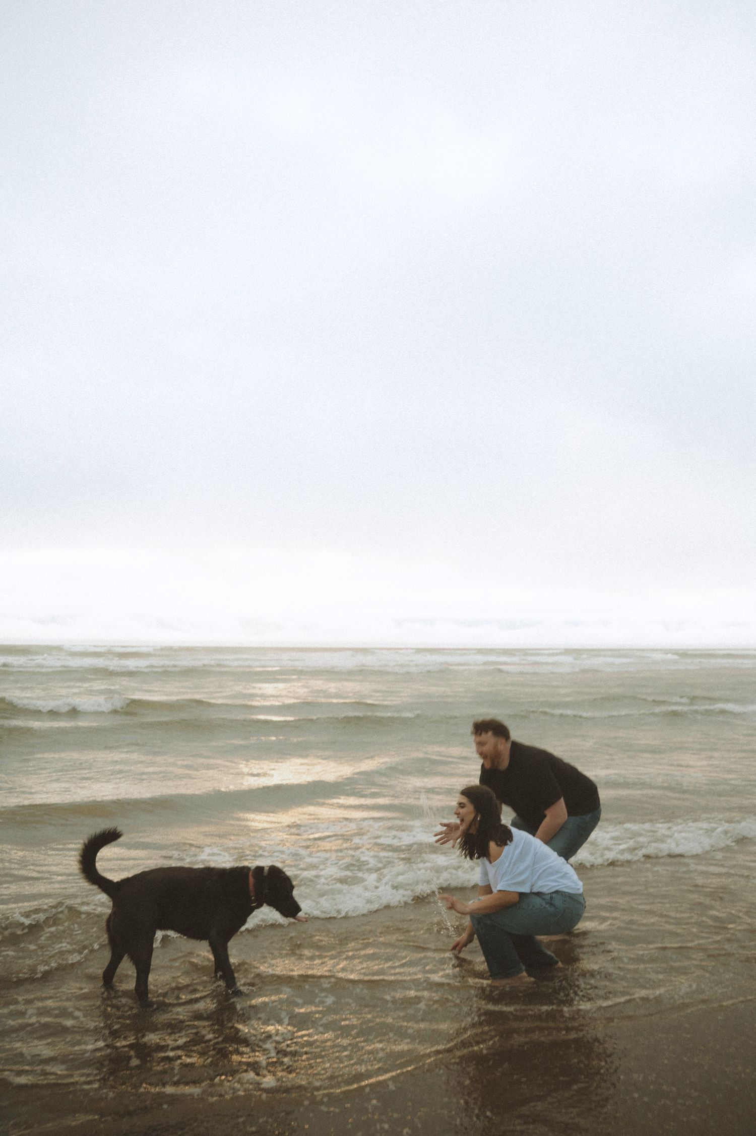 Couple playing with black Labrador dog on a cloudy beach, waves washing over their feet at the shoreline.