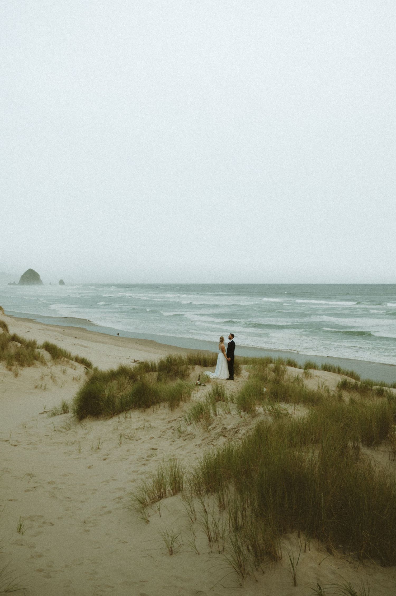 Two figures stand on coastal dunes overlooking a misty Pacific Ocean beach.