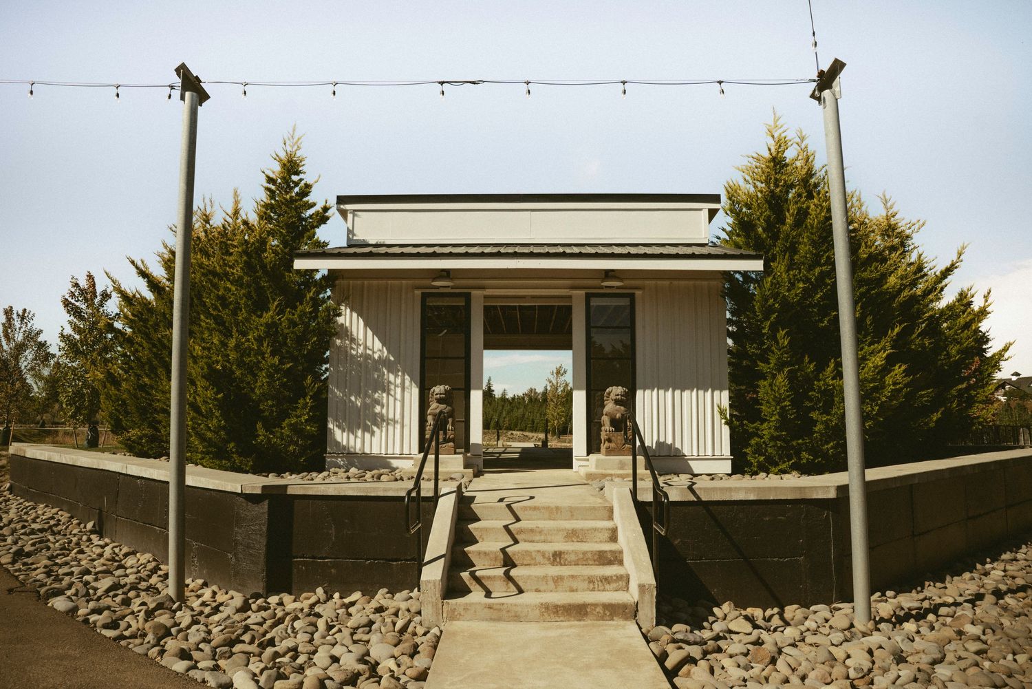 Small modern building with stone steps, flanked by tall evergreen trees and decorative statues, under a clear sky.