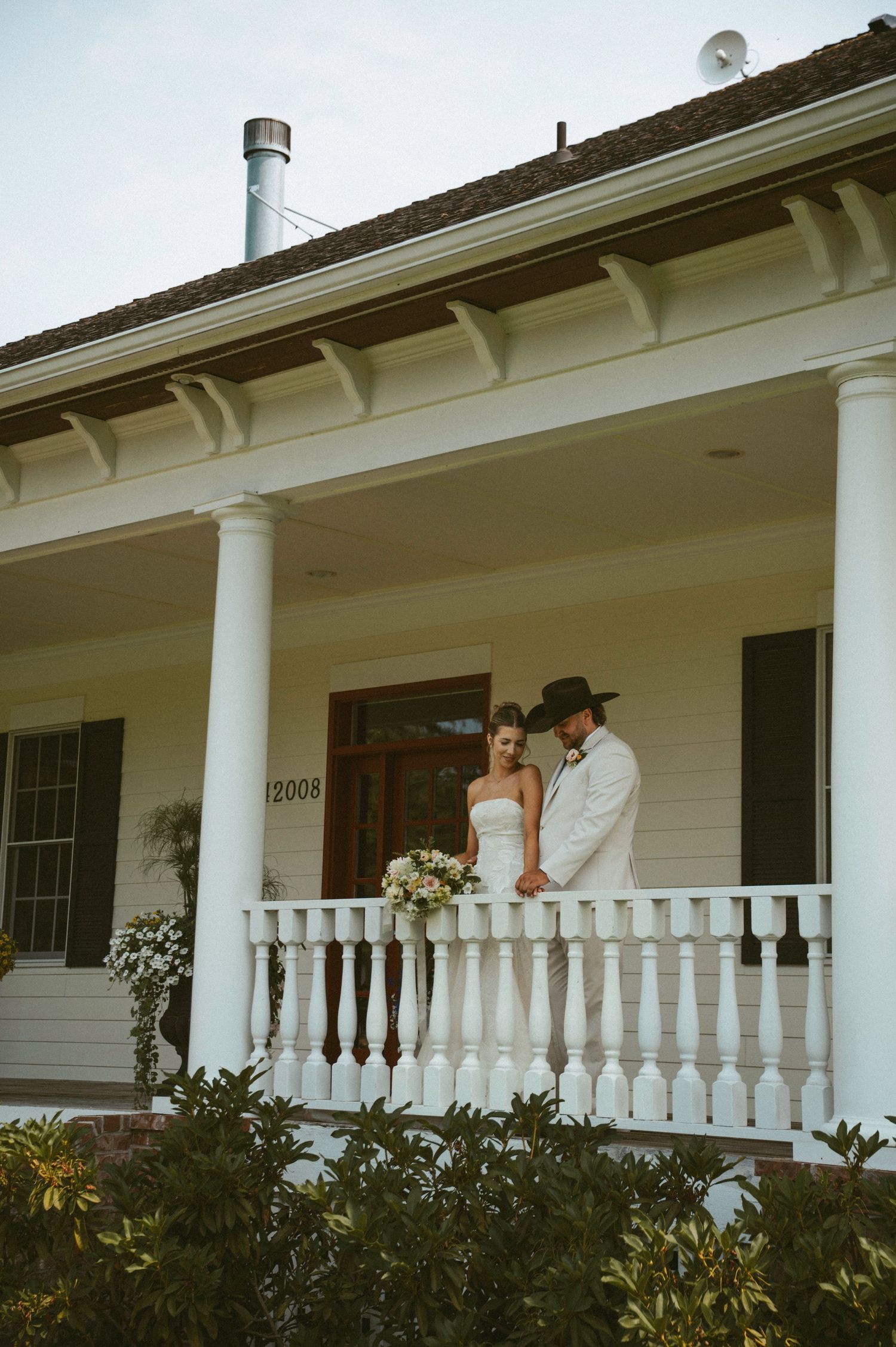 Bride and groom in white attire stand on a charming white porch with columns at house number 12008.