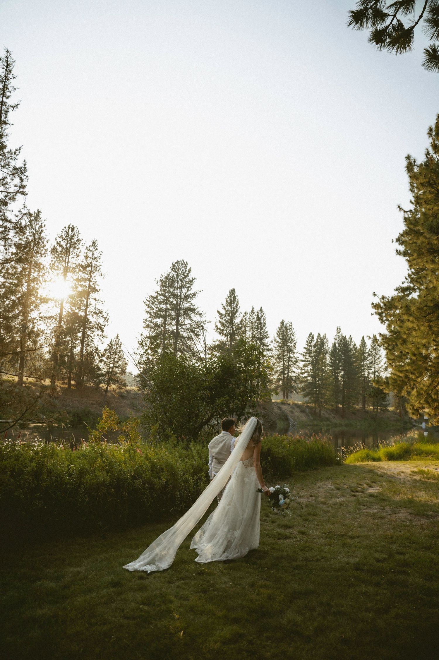 Bride and groom embrace at golden hour sunset in a lush forest meadow, wedding veil trailing gracefully on green grass.