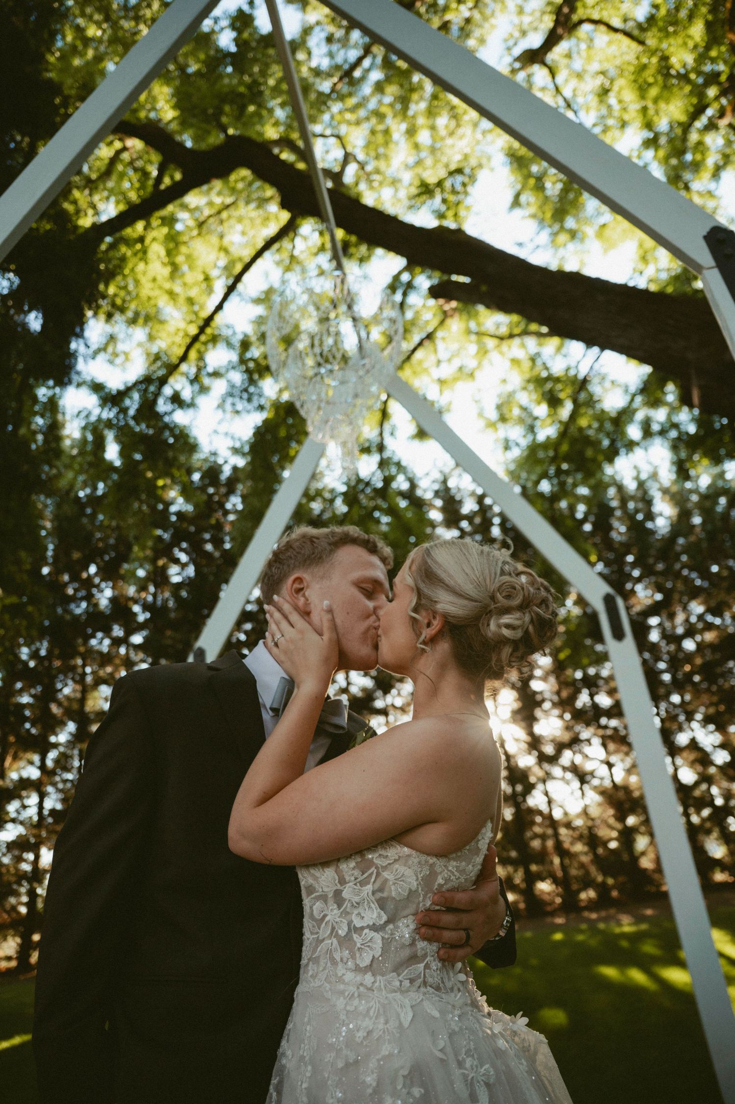 Bride and groom share first kiss under white arch surrounded by lush green trees at outdoor wedding ceremony.