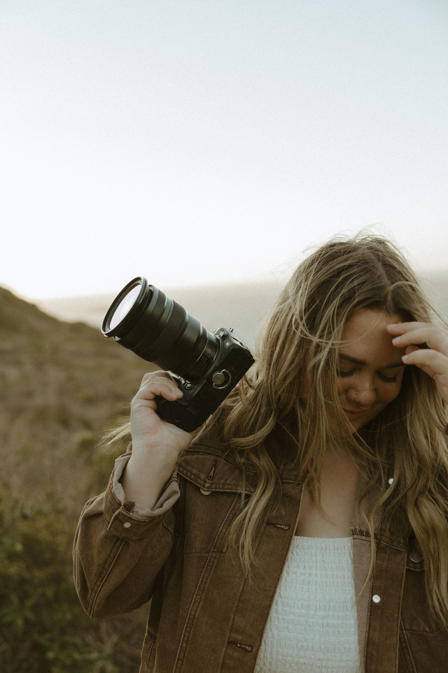 Person in brown jacket holding camera against muted natural background.
