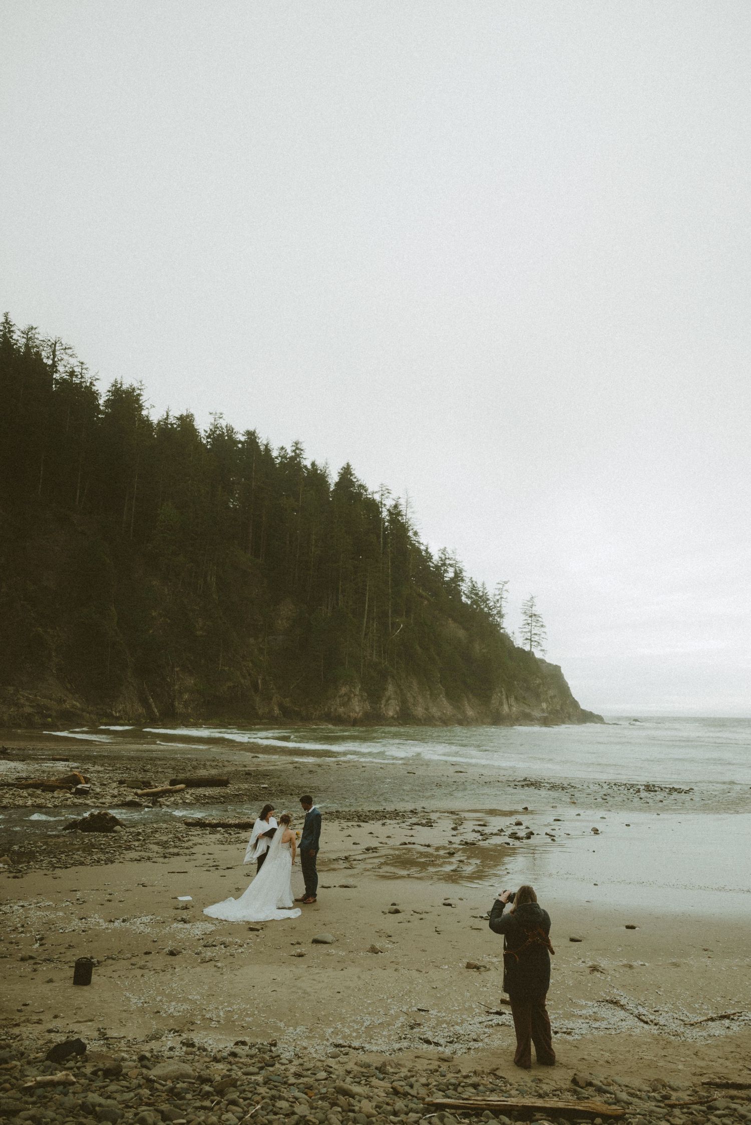 A couple poses for wedding photos on a misty beach with towering evergreen cliffs in the Pacific Northwest.